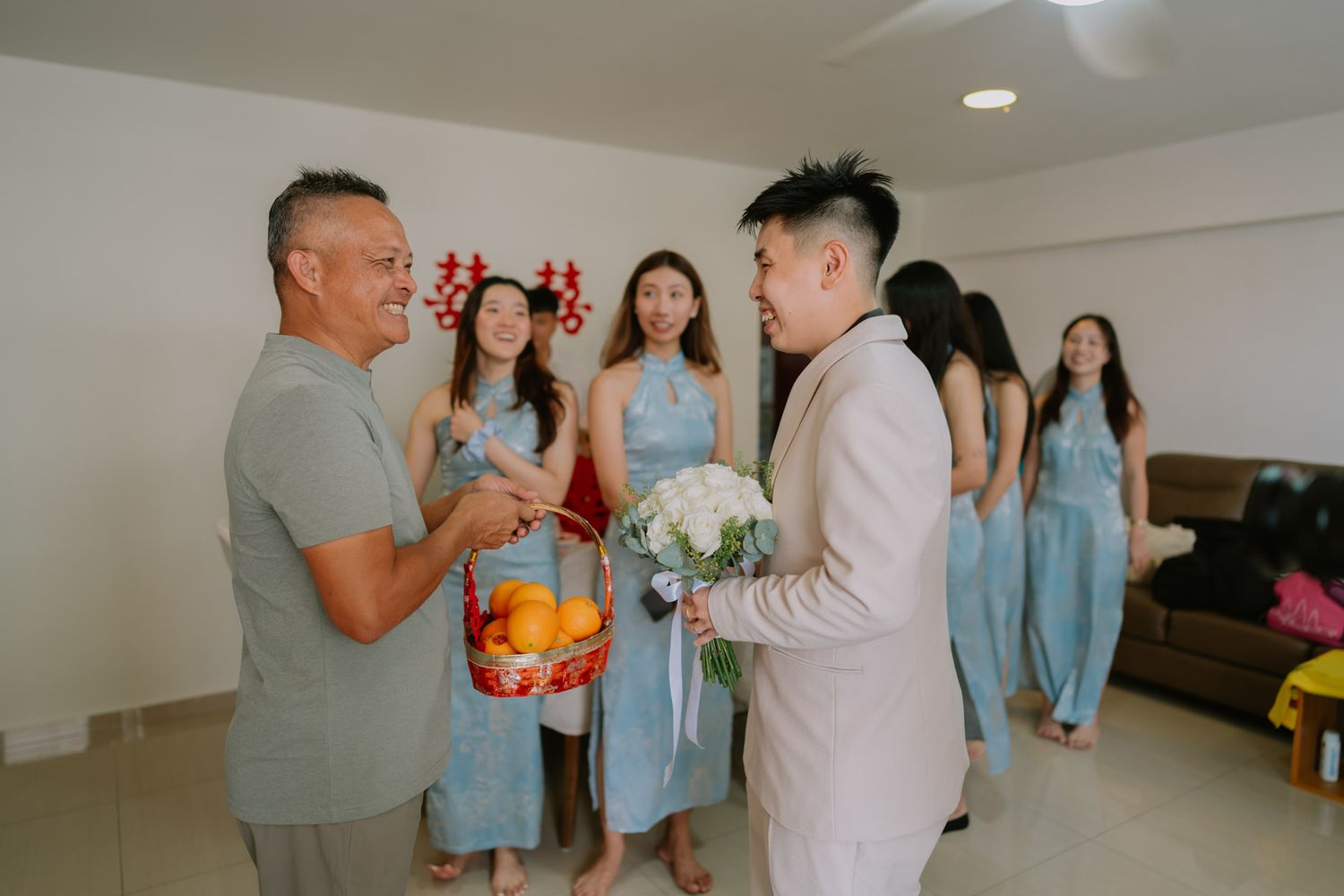 Groom in white suit presents bouquet to family during Asian wedding door game ceremony with bridesmaids in blue dresses.