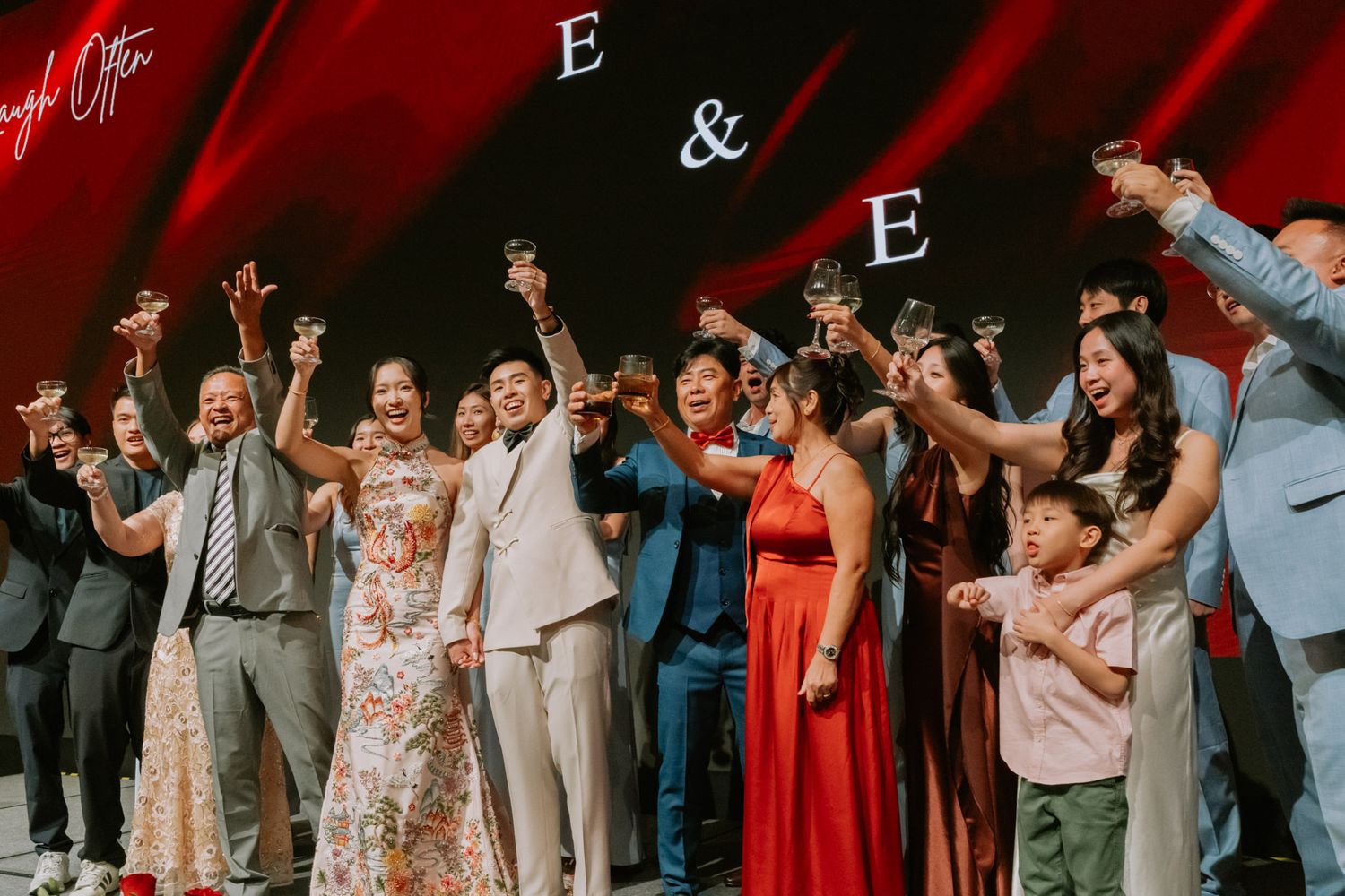 Joyful wedding reception toast with guests raising glasses in front of a red backdrop with monogram letters E and E.