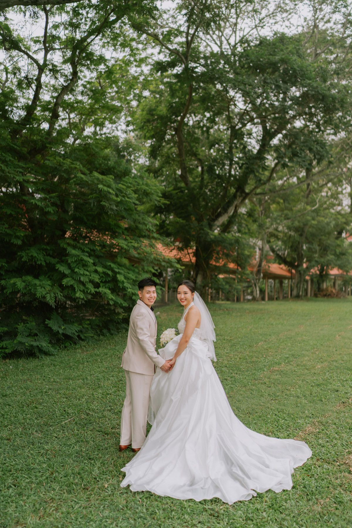 Bride and groom holding hands in lush green park, bride in white gown with train, groom in beige suit.