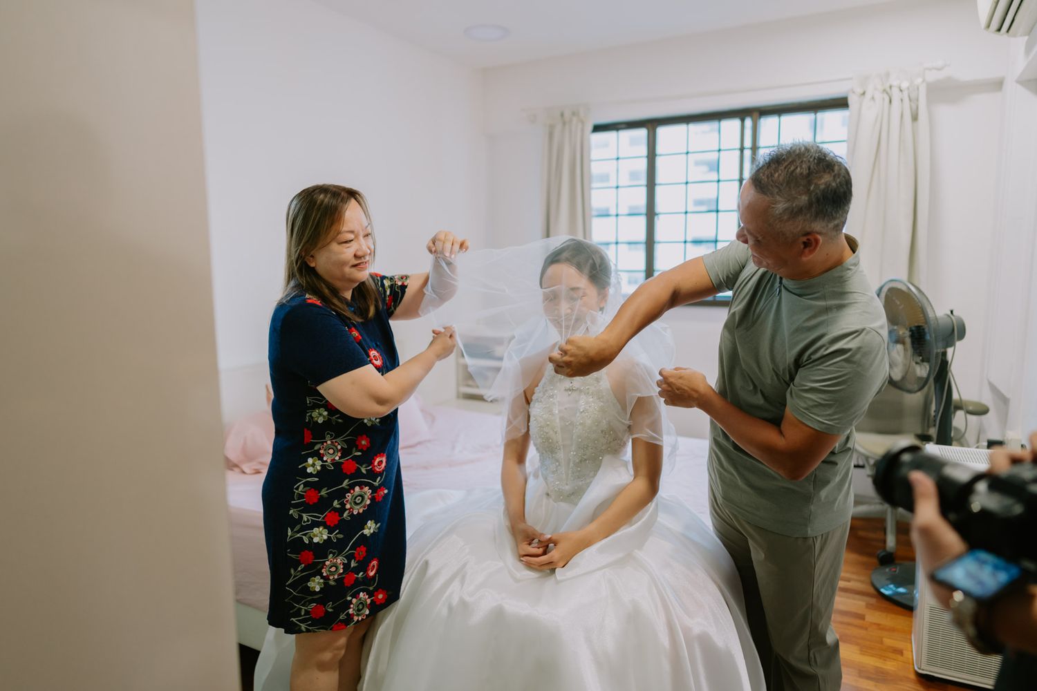 Bride in white wedding gown being helped by two people adjusting her veil in a bright indoor room.