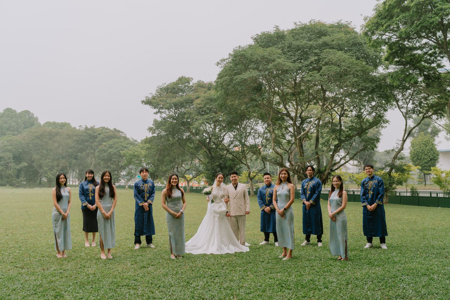 Wedding party poses on lush green lawn, bride and groom centered among bridesmaids and groomsmen in blue-gray attire.