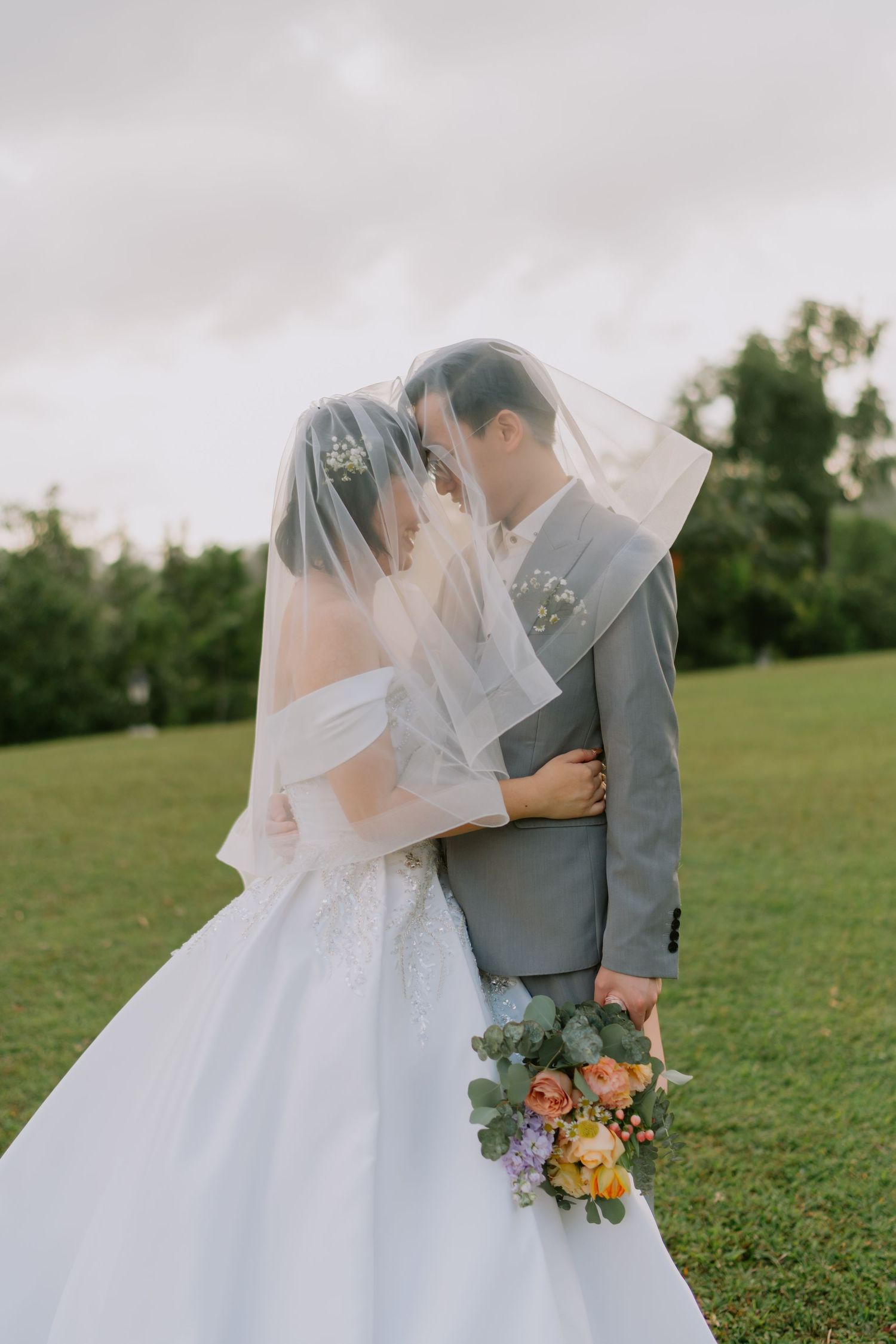 Bride and groom share an intimate moment under the bridal veil on a green lawn outdoors.