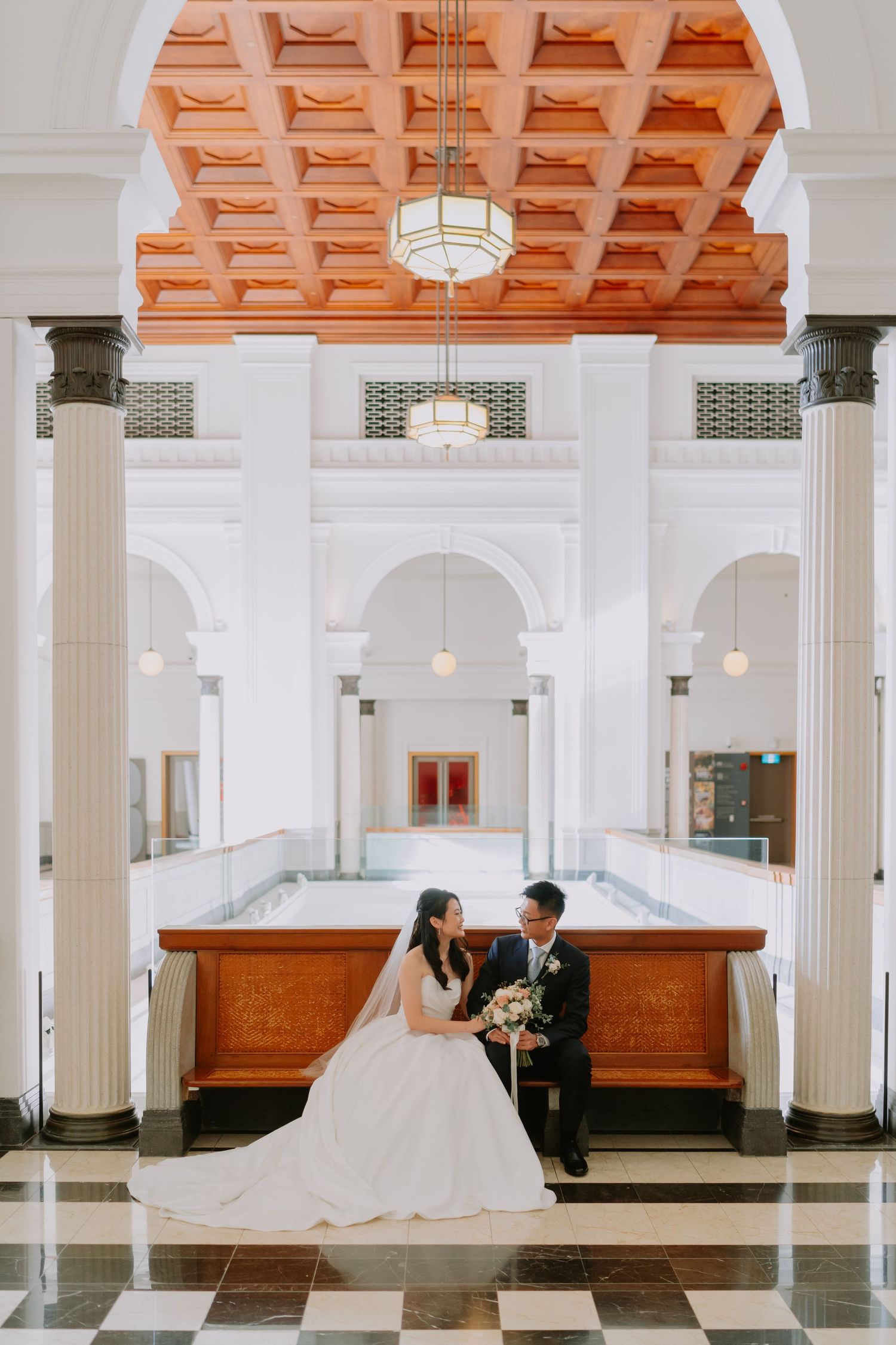 Wedding couple seated together at wooden counter beneath ornate coffered ceiling and columns.
