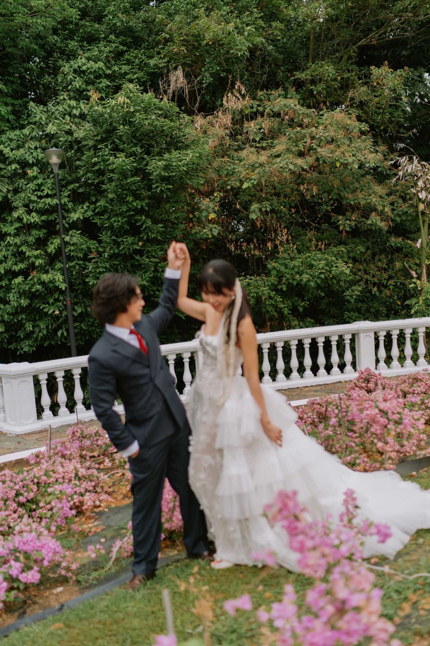 Groom spinning bride in a tiered white gown amid pink blooming flowers near a white garden balustrade.