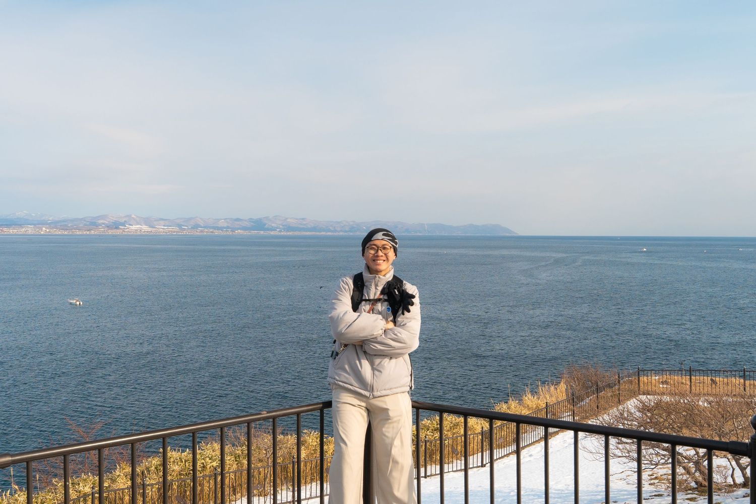 Person standing on snowy balcony overlooking calm blue sea on a clear winter day.