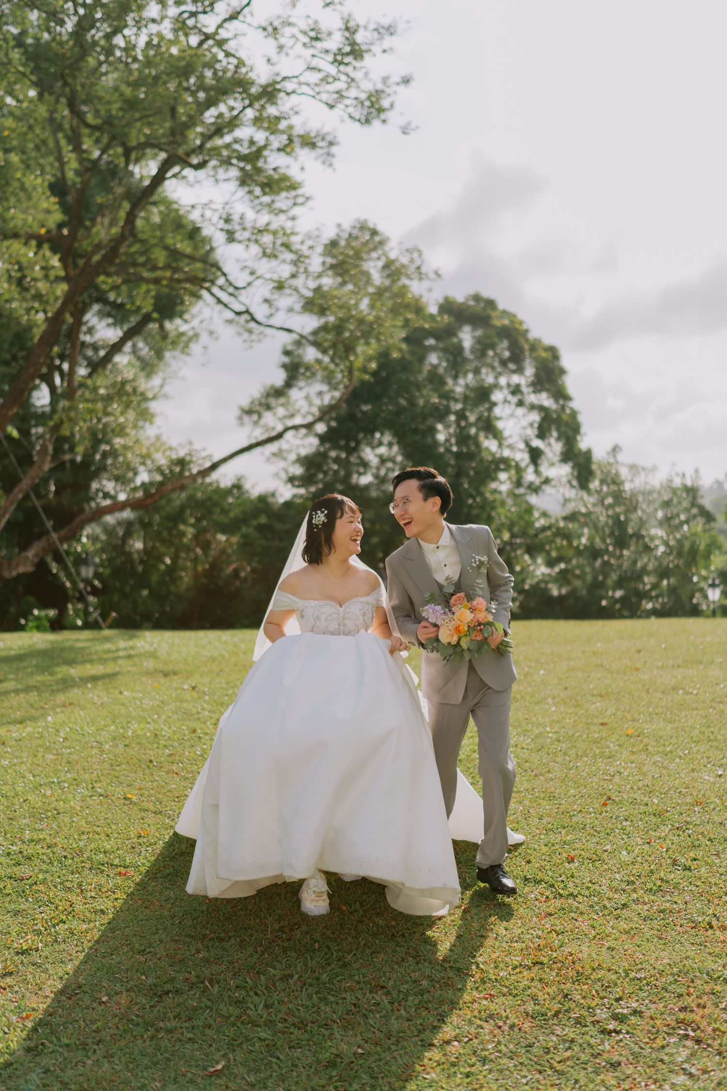 Smiling bride and groom walk together on a sunny green lawn holding a colorful wedding bouquet.