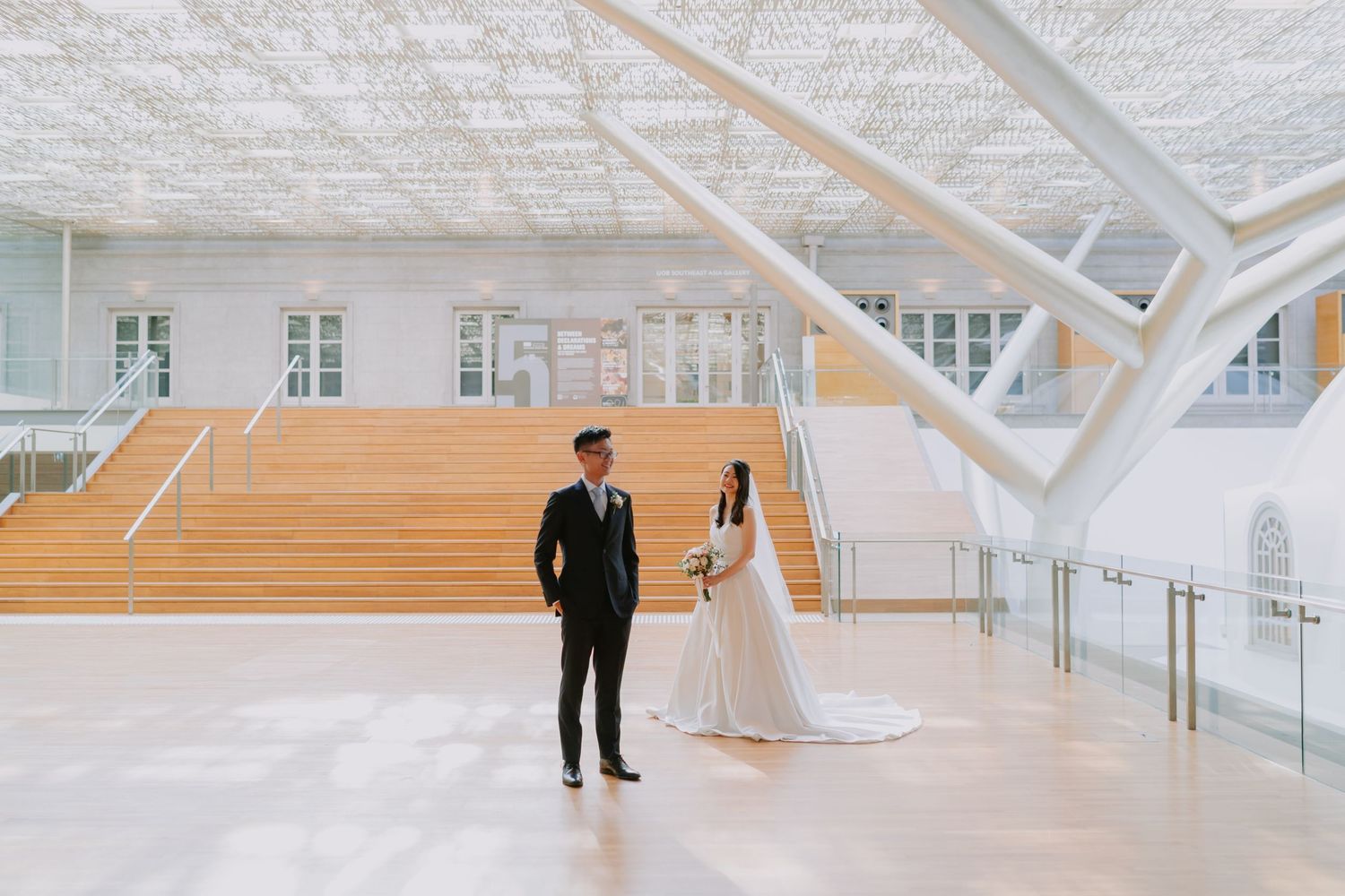 Bride and groom standing apart in modern white architectural wedding venue with geometric ceiling and grand staircase.