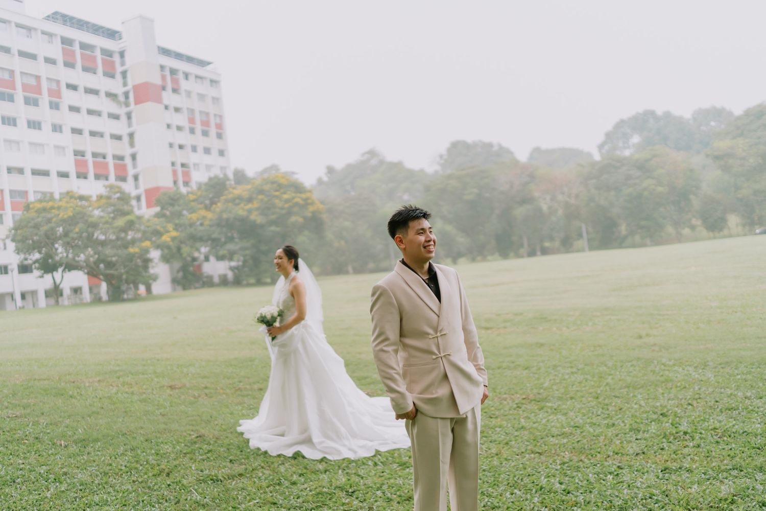 A groom in a beige suit smiles while his bride in a white gown holds flowers on a lush green field.