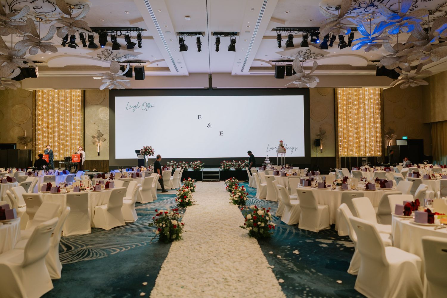 Elegant wedding banquet hall with white chair covers, floral aisle, large projection screen, and decorative ceiling at a luxury venue.