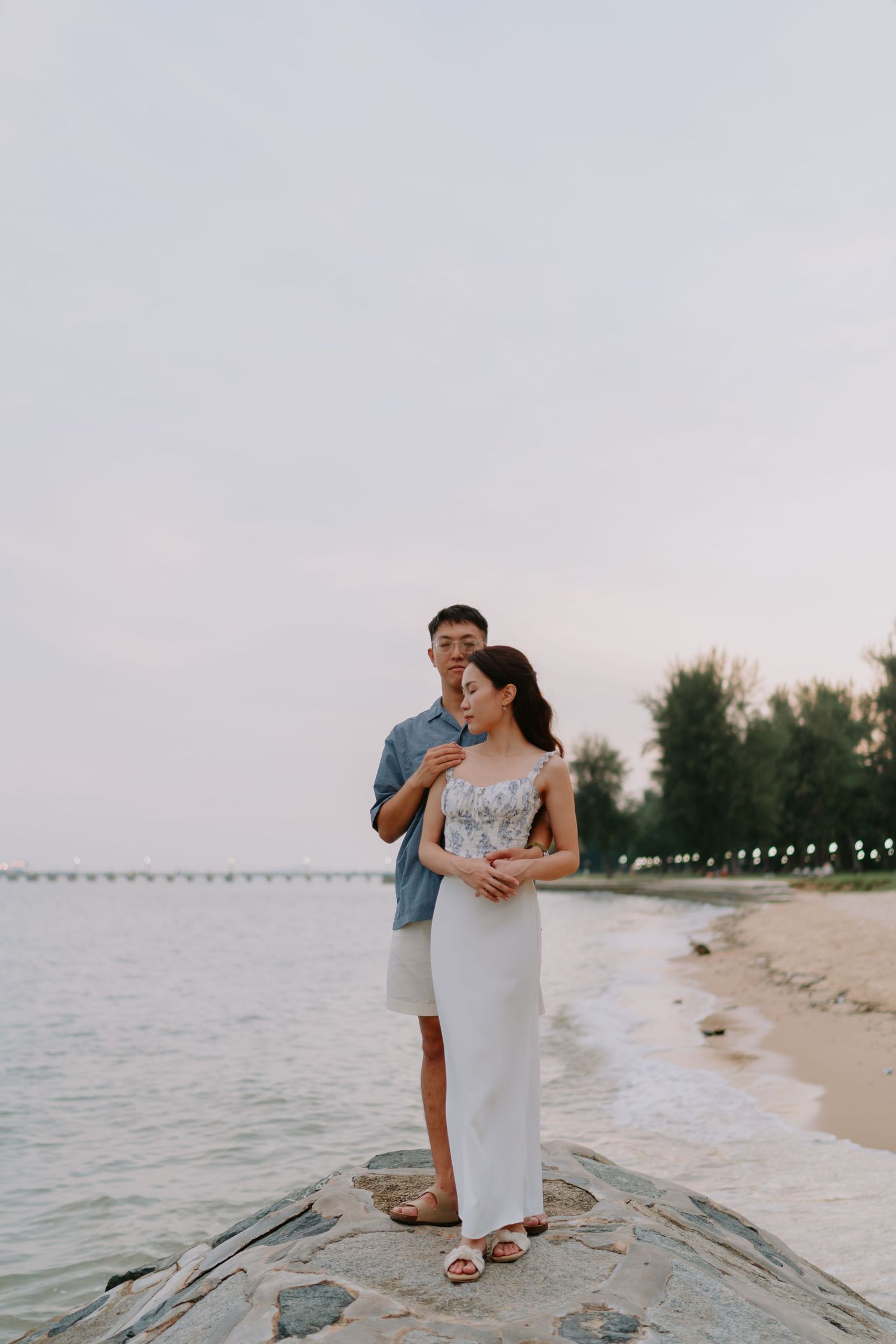 Couple standing on coastal rocks embracing at sunset with calm sea and bridge behind.