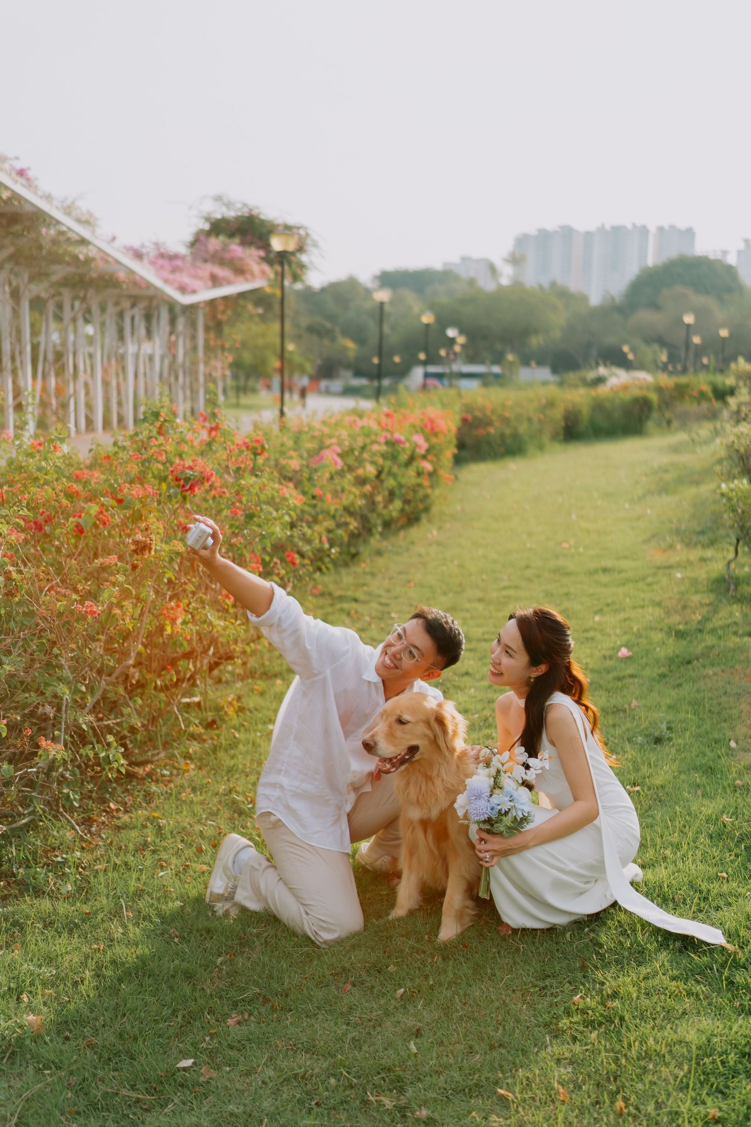 Couple taking selfie with golden retriever in a colorful flower garden at sunset.