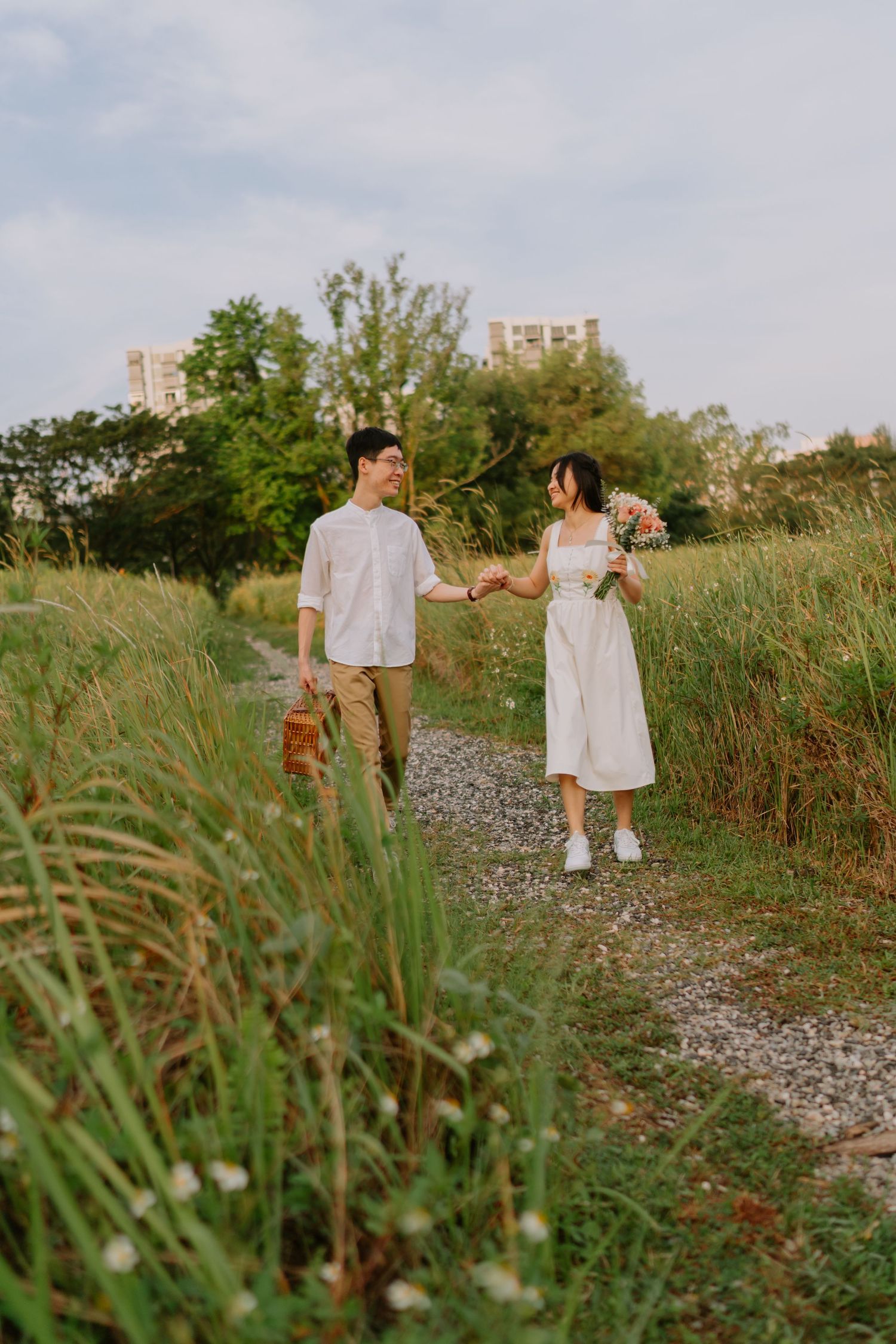 Couple holding hands on a gravel path through tall grass, woman in white dress holding bouquet.