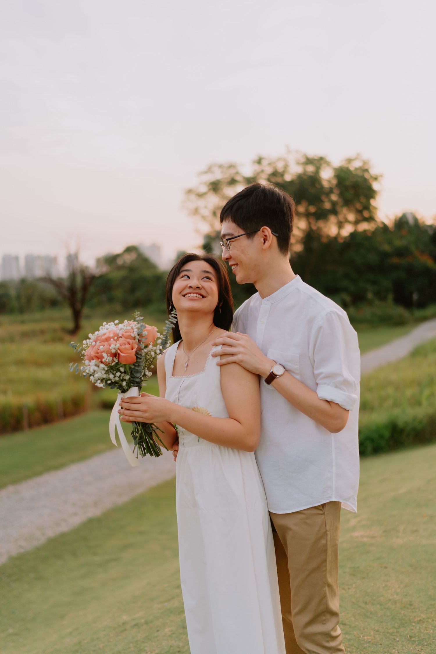 A smiling couple stands together on a green lawn at sunset, the woman holding a floral bouquet.