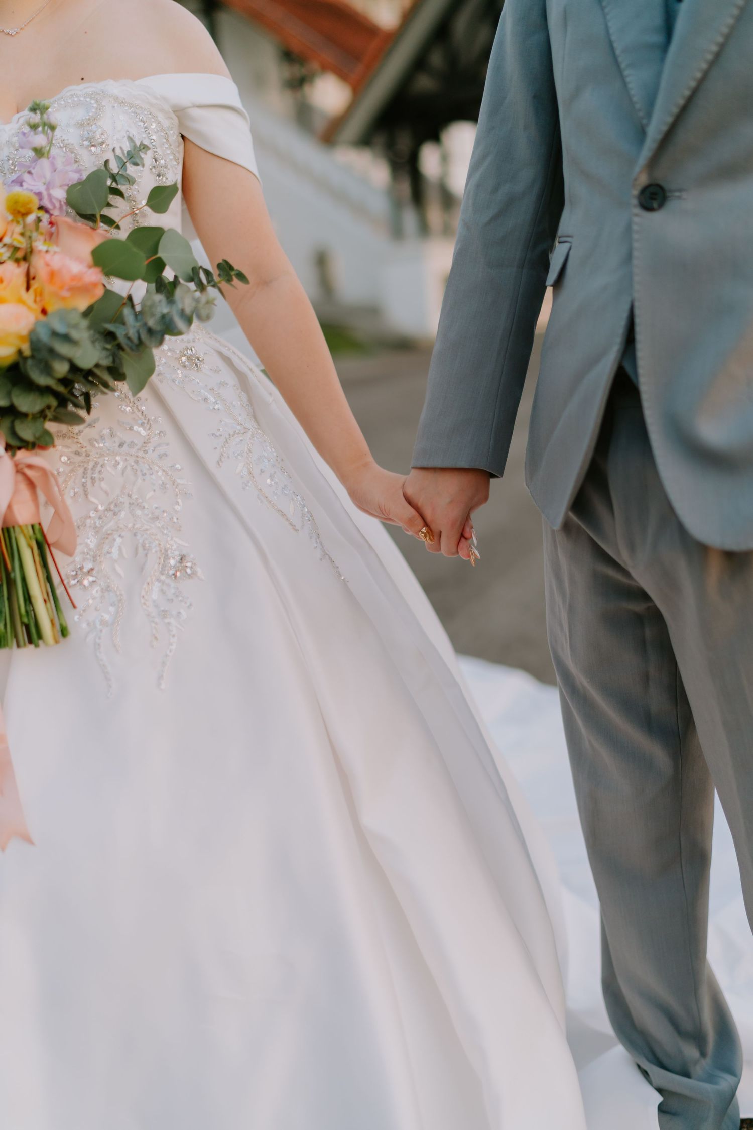 Bride and groom holding hands, bride in white off-shoulder gown with floral bouquet, groom in grey suit.