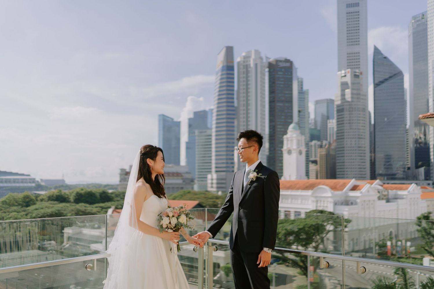 Bride and groom holding hands on a rooftop balcony with Singapore skyline in the background.