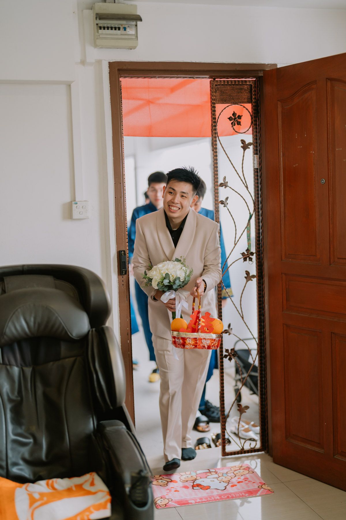 Smiling groom in beige suit holding white bouquet and red gift basket entering home doorway at Asian wedding ceremony.