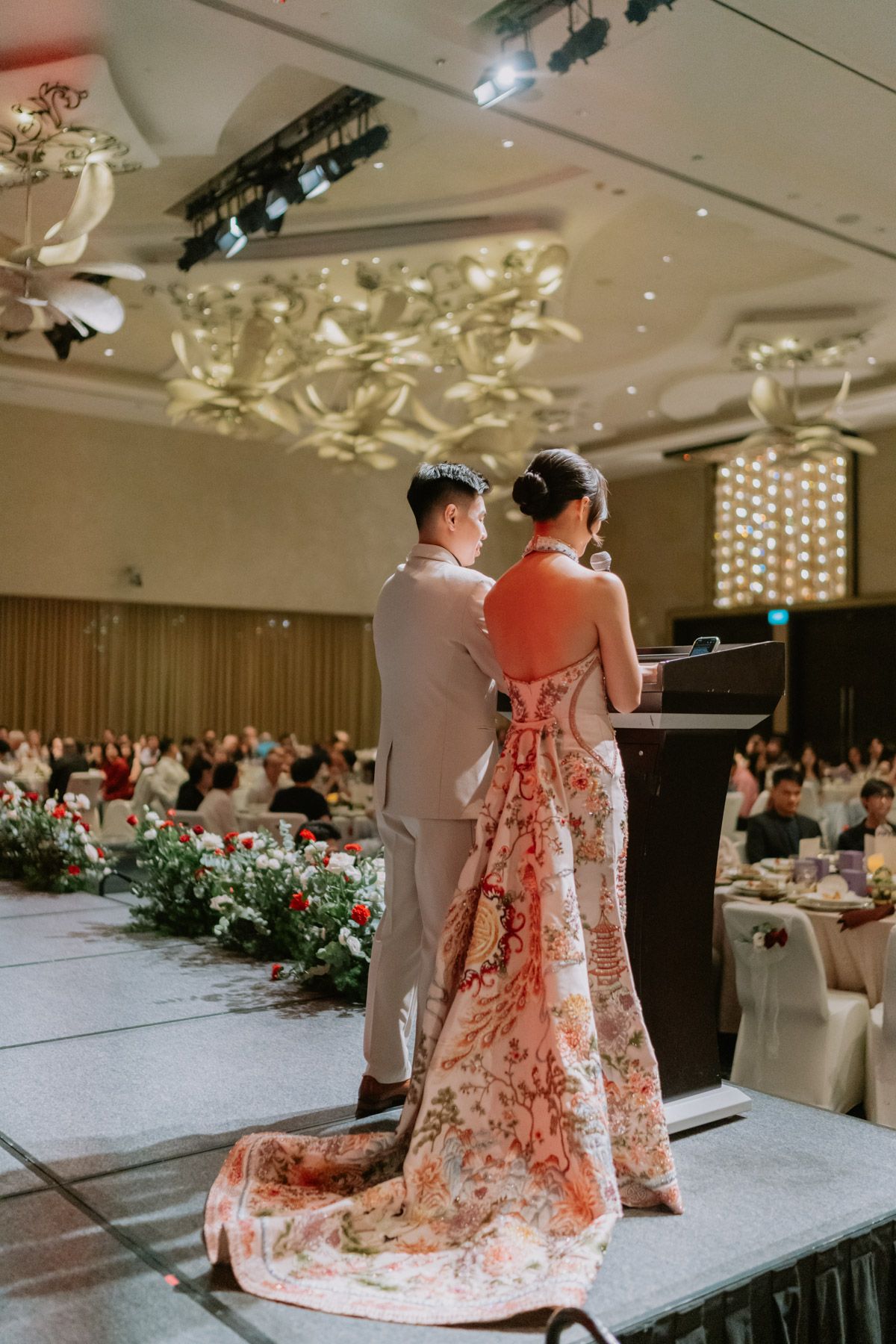 Couple in elegant wedding attire standing at podium in grand ballroom during reception ceremony.