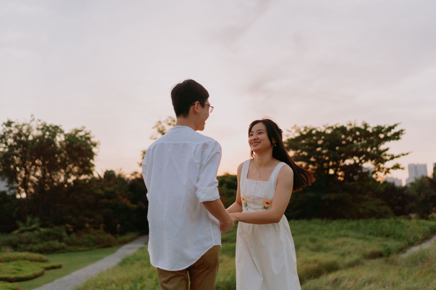A couple holds hands facing each other on a grassy path at sunset in a lush green park.