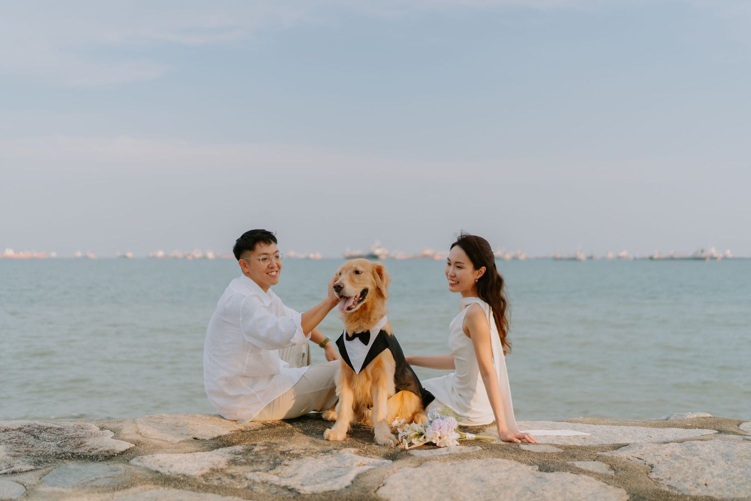 Couple sitting on rocks by the sea with their golden retriever dressed in a tuxedo.