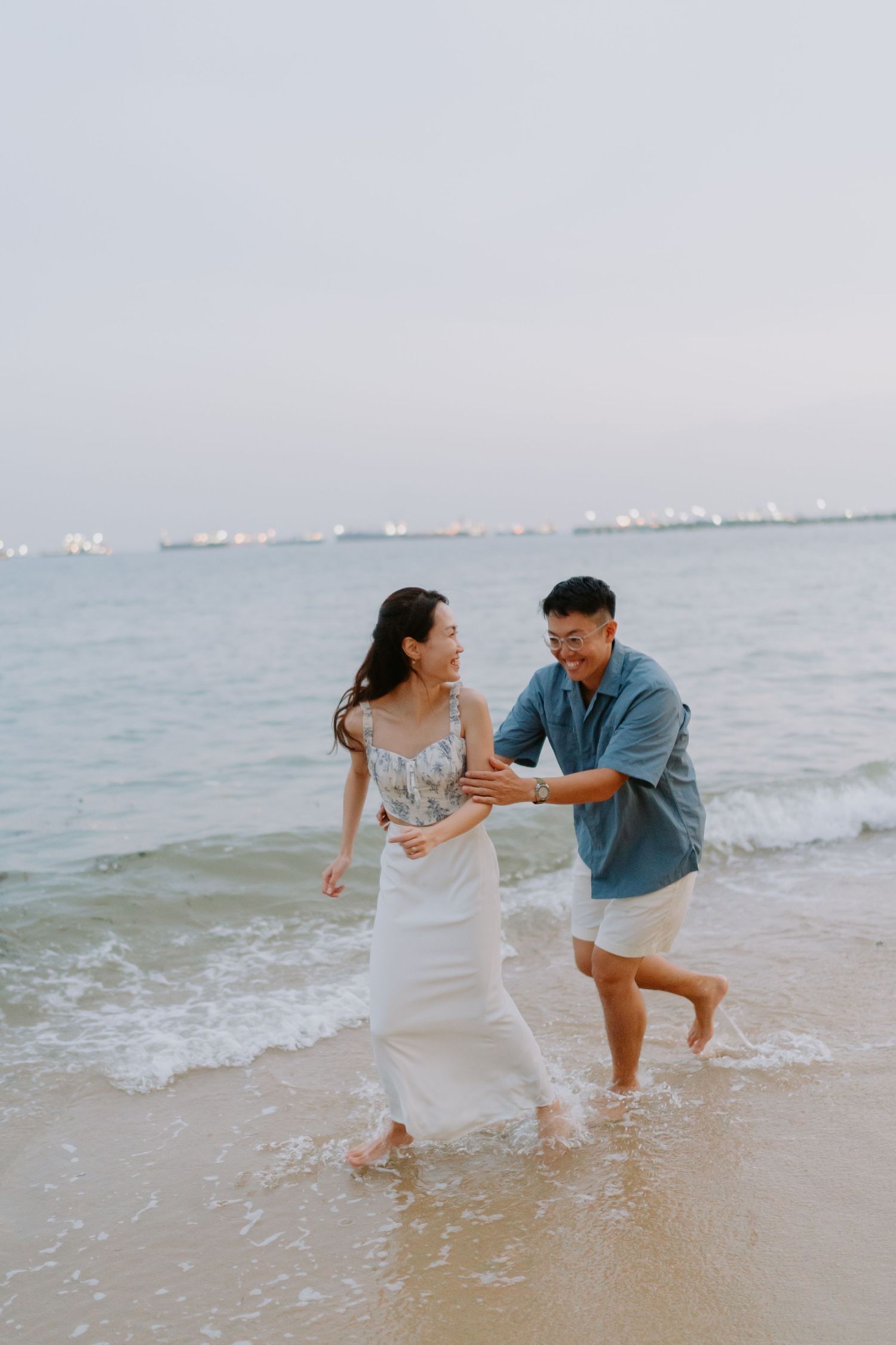 Joyful couple laughing and playing together in ocean waves on sandy beach at dusk.