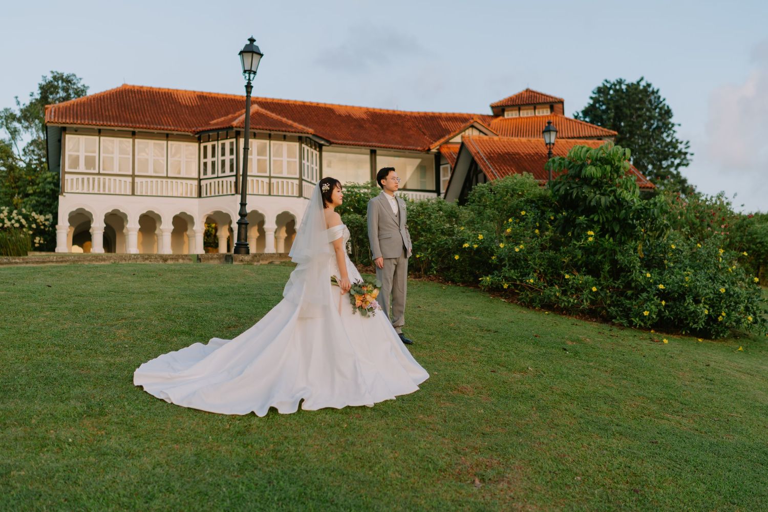 Bride and groom posing on green lawn before elegant colonial building at golden hour sunset.