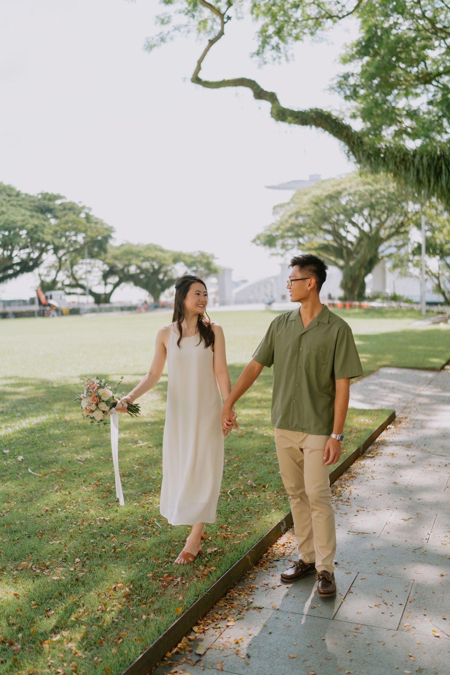 Couple holding hands walking on a path in a sunlit park with bouquet.