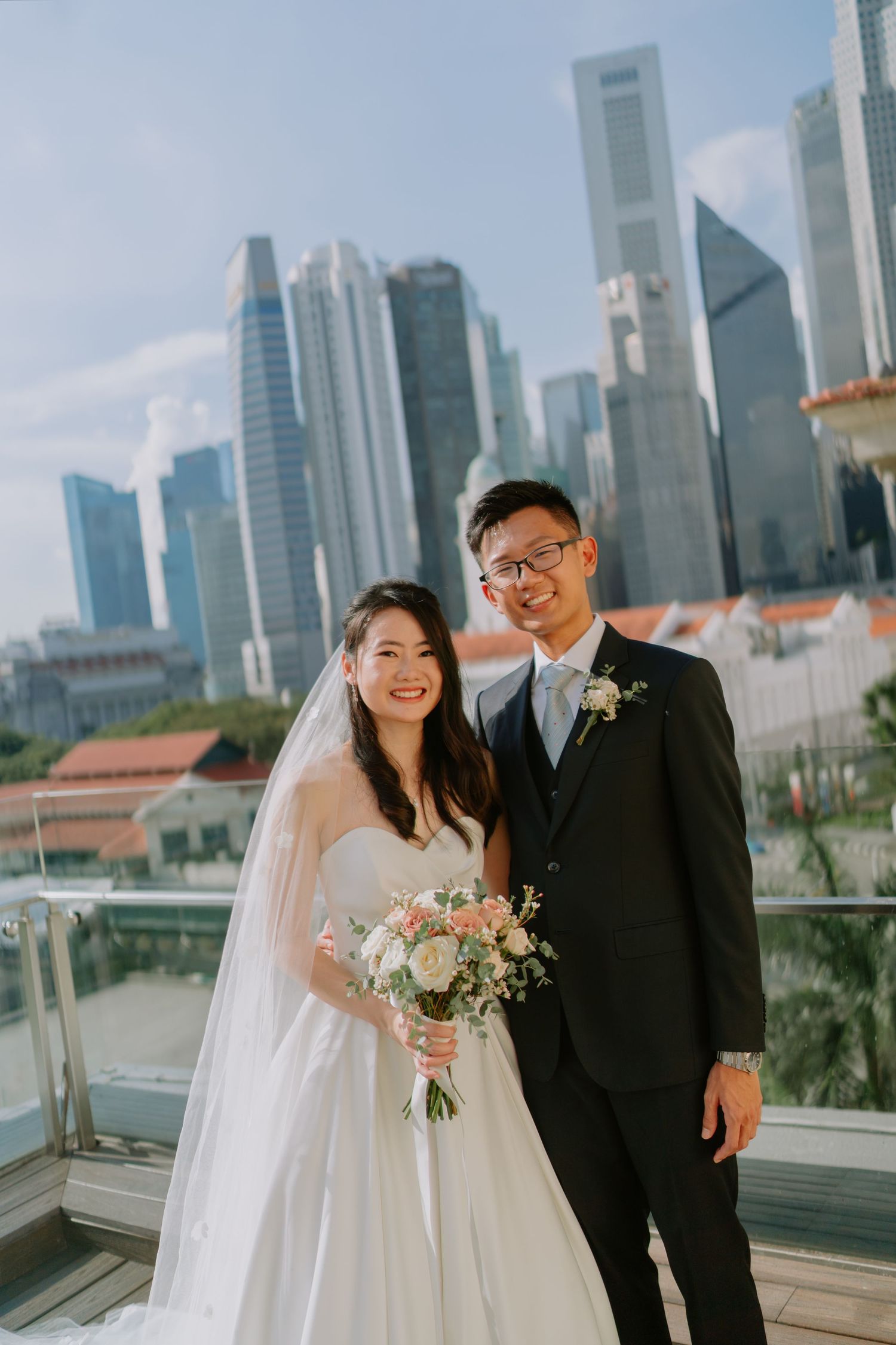 Smiling bride and groom pose together on rooftop with Singapore city skyline behind them.
