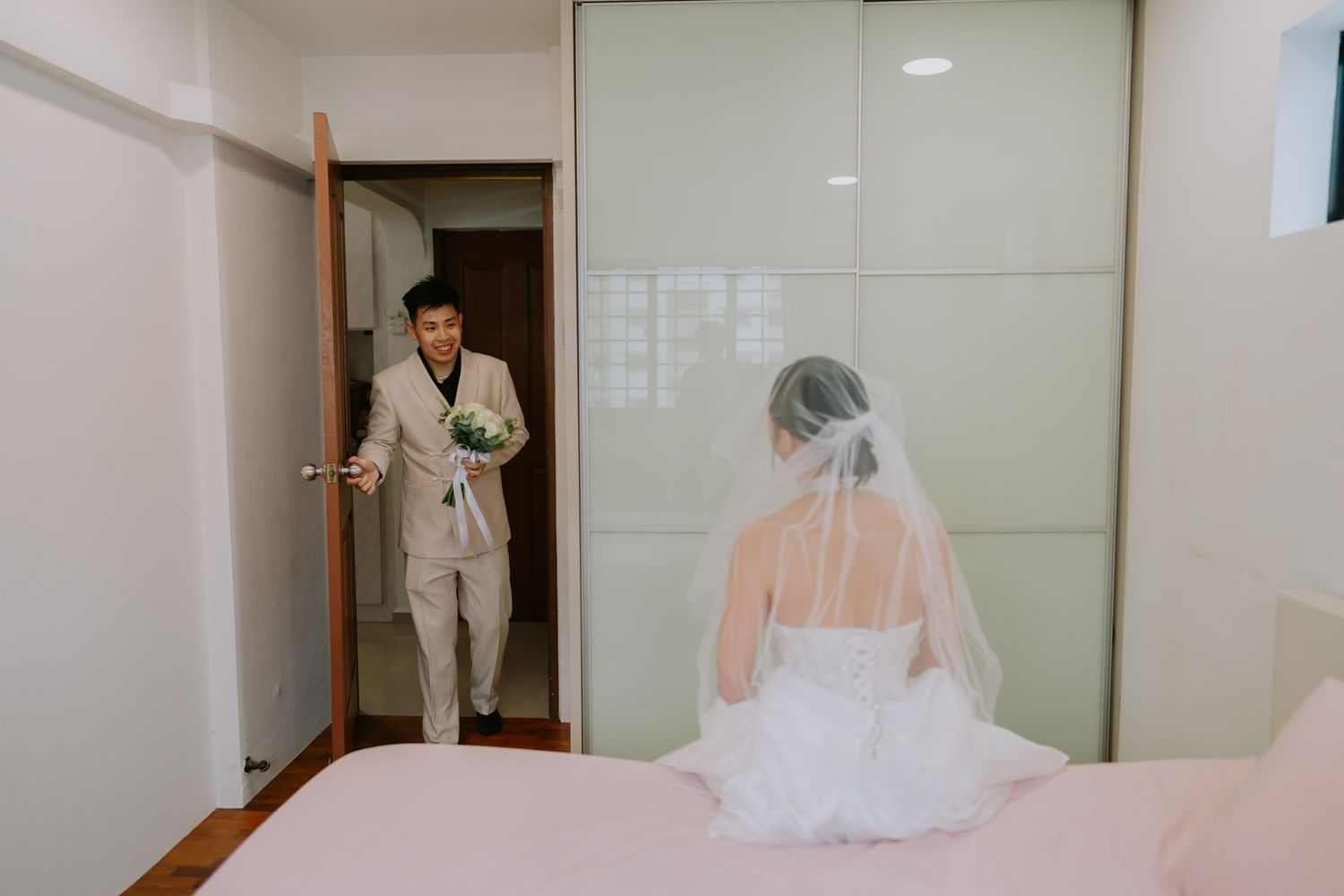 Groom in beige suit entering bedroom holding bouquet, bride in white wedding gown and veil seated with back turned.