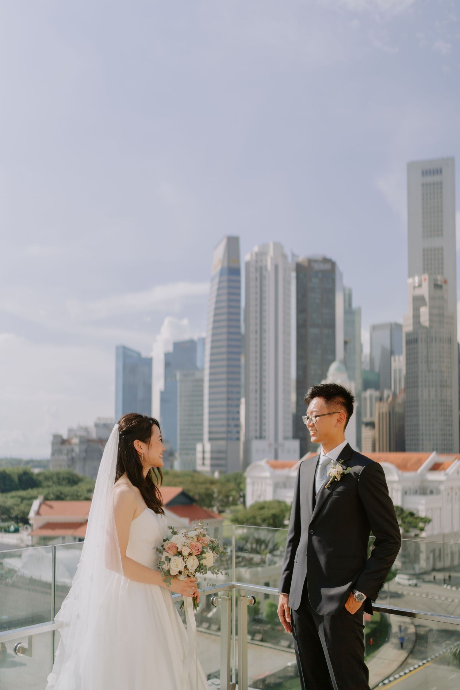 Bride and groom standing on a rooftop terrace with the Singapore city skyline behind them.