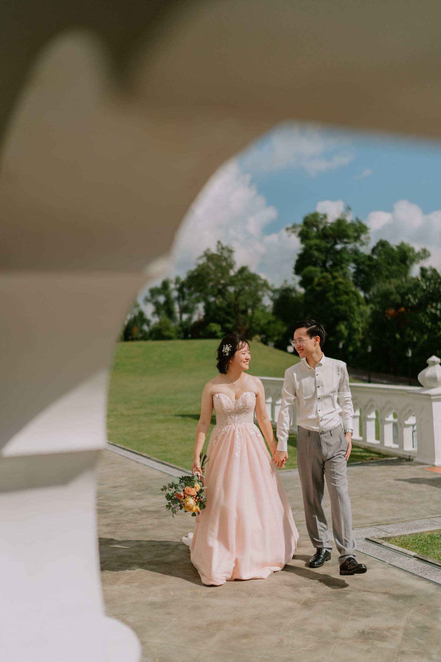 Bride in blush pink gown and groom in grey suit walk hand in hand on sunlit outdoor terrace.