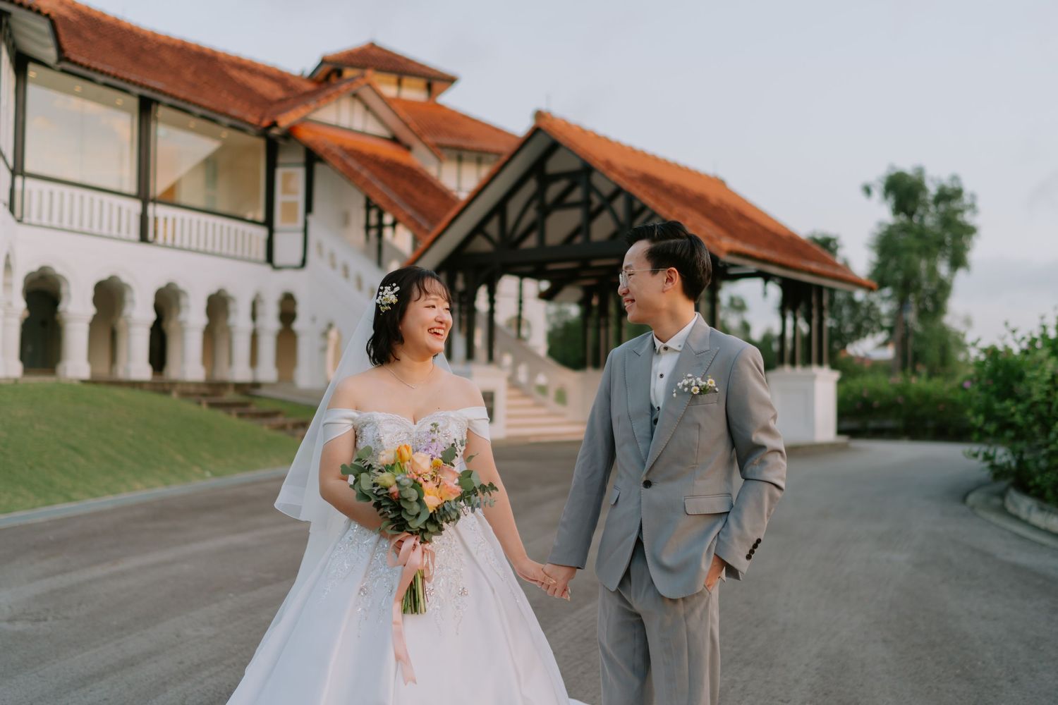 A bride and groom walk hand in hand smiling near a beautiful historic colonial venue.