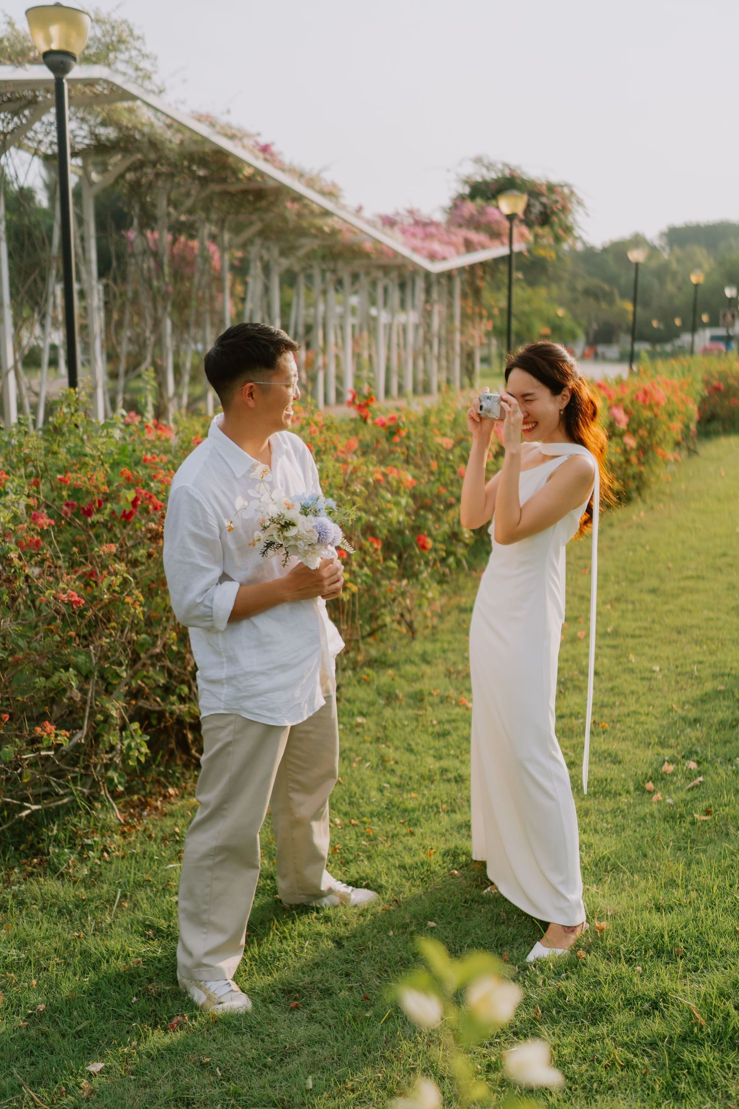 Woman in white dress photographing man holding bouquet near flowering garden pergola.