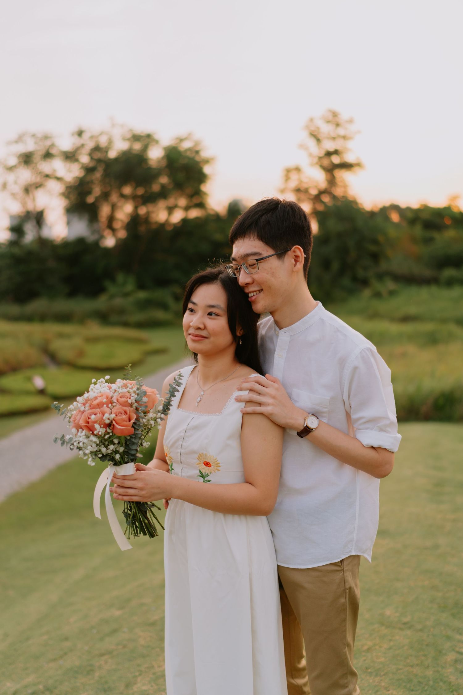 A couple embraces at sunset in a park, the woman holding a bouquet of pink and white flowers.