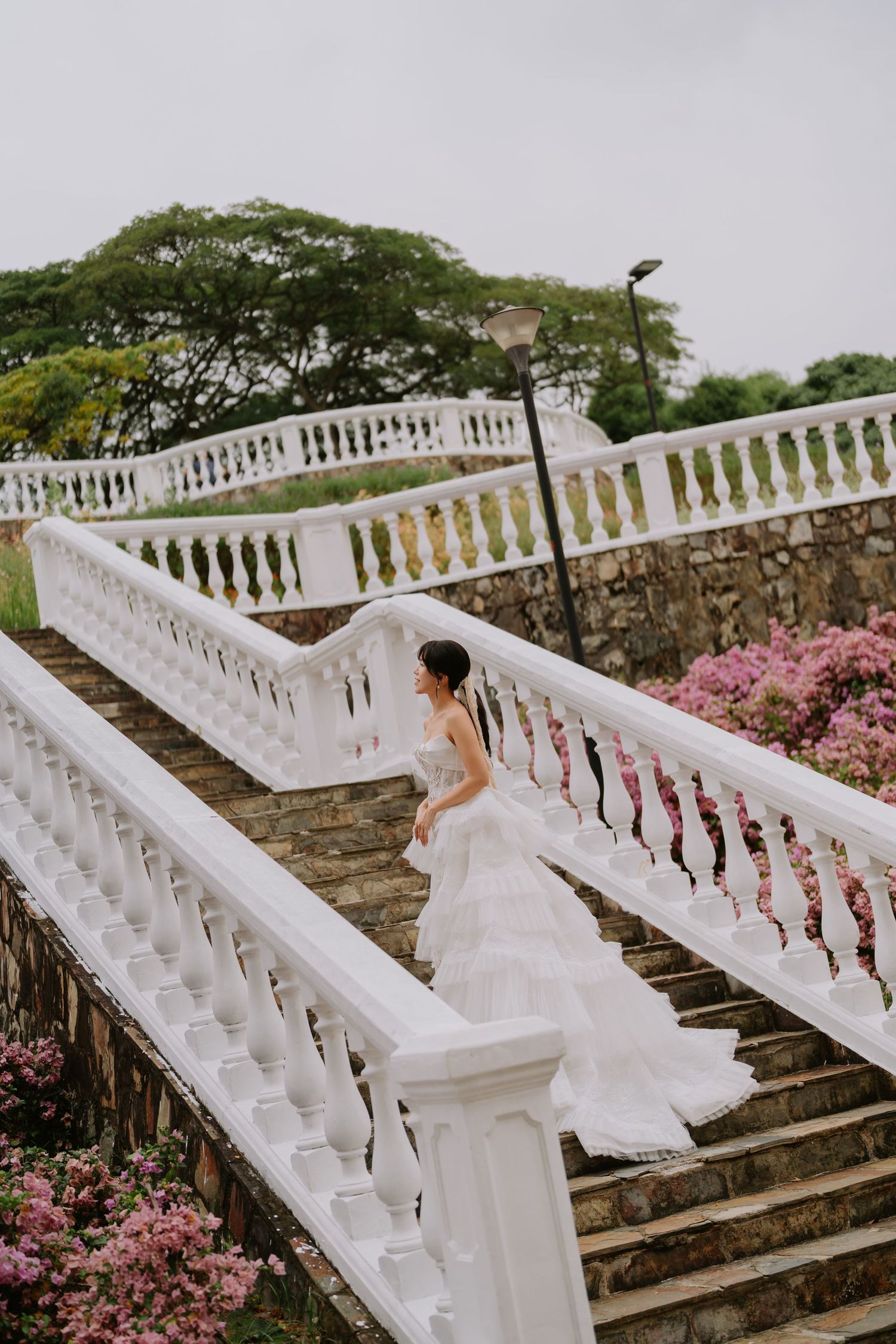 Elegant bride in white tiered gown ascending white balustrade stairs surrounded by pink flowers.
