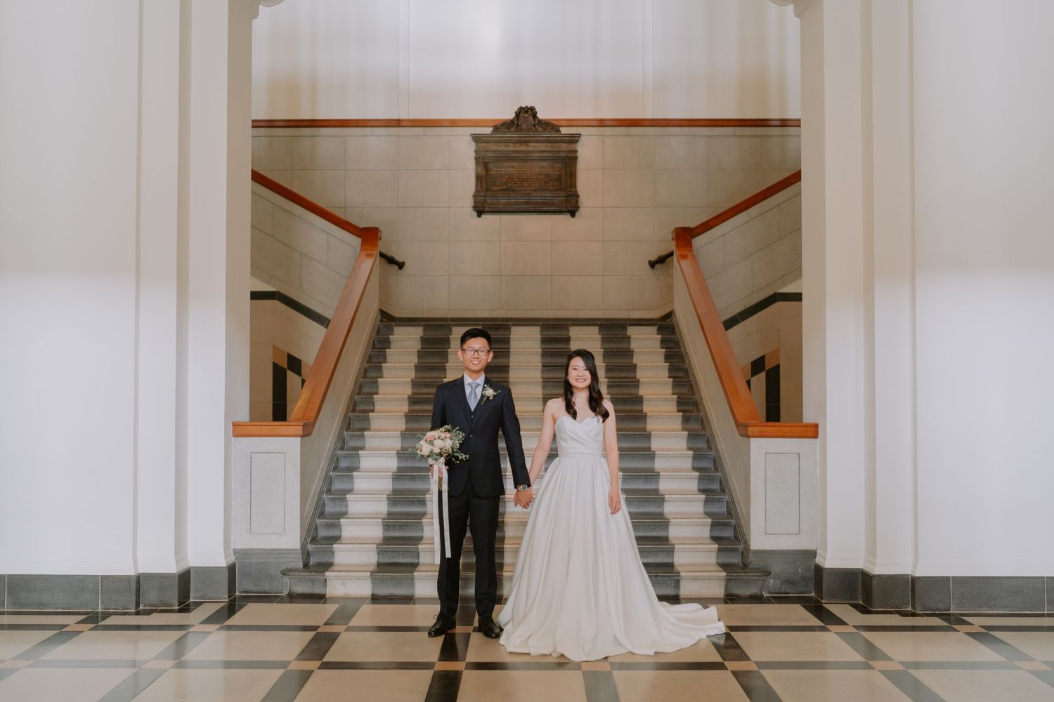 Bride and groom stand before grand staircase in classic marble foyer with checkered tile floor.