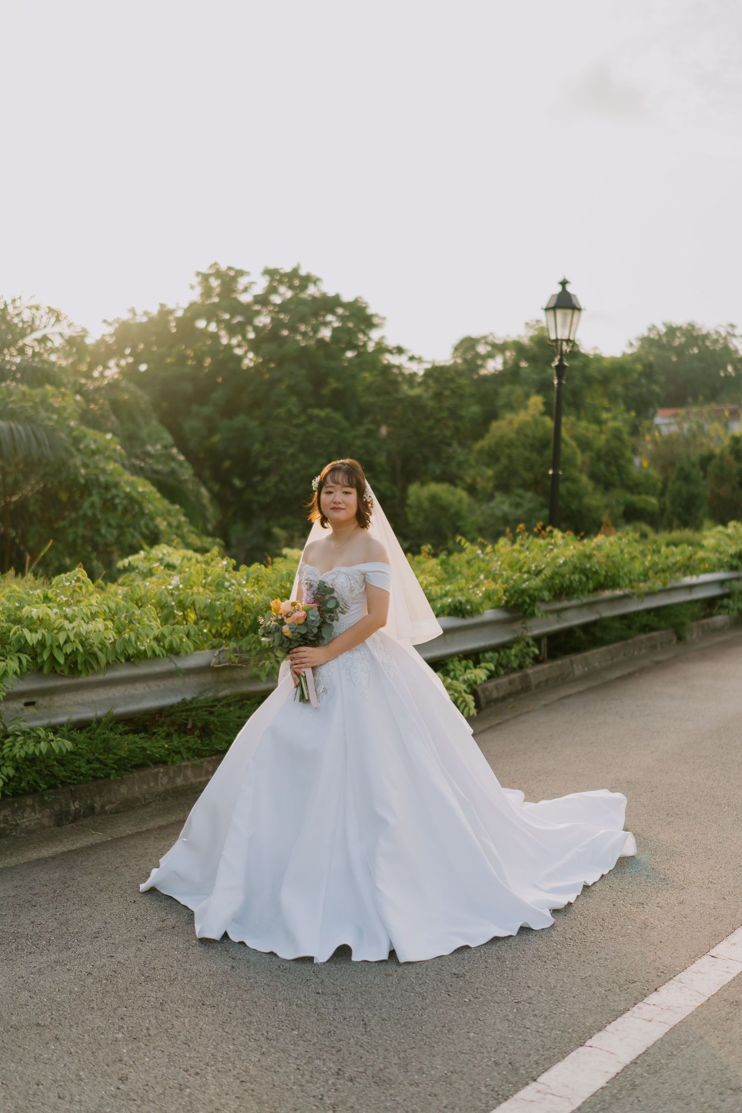 Bride in off-shoulder white ballgown with long train holds bouquet on road at golden sunset.