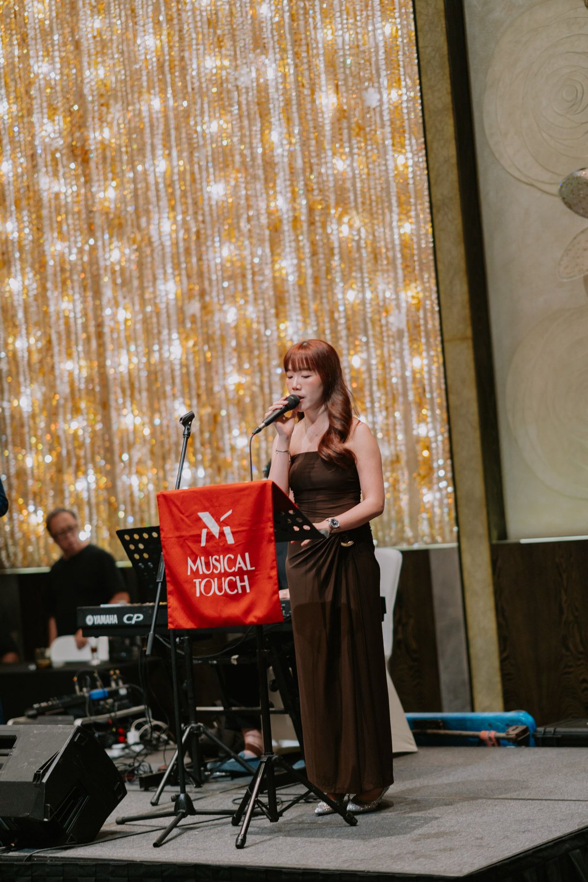A woman in a brown gown sings on stage at a Musical Touch event, backed by a sparkling gold curtain backdrop.