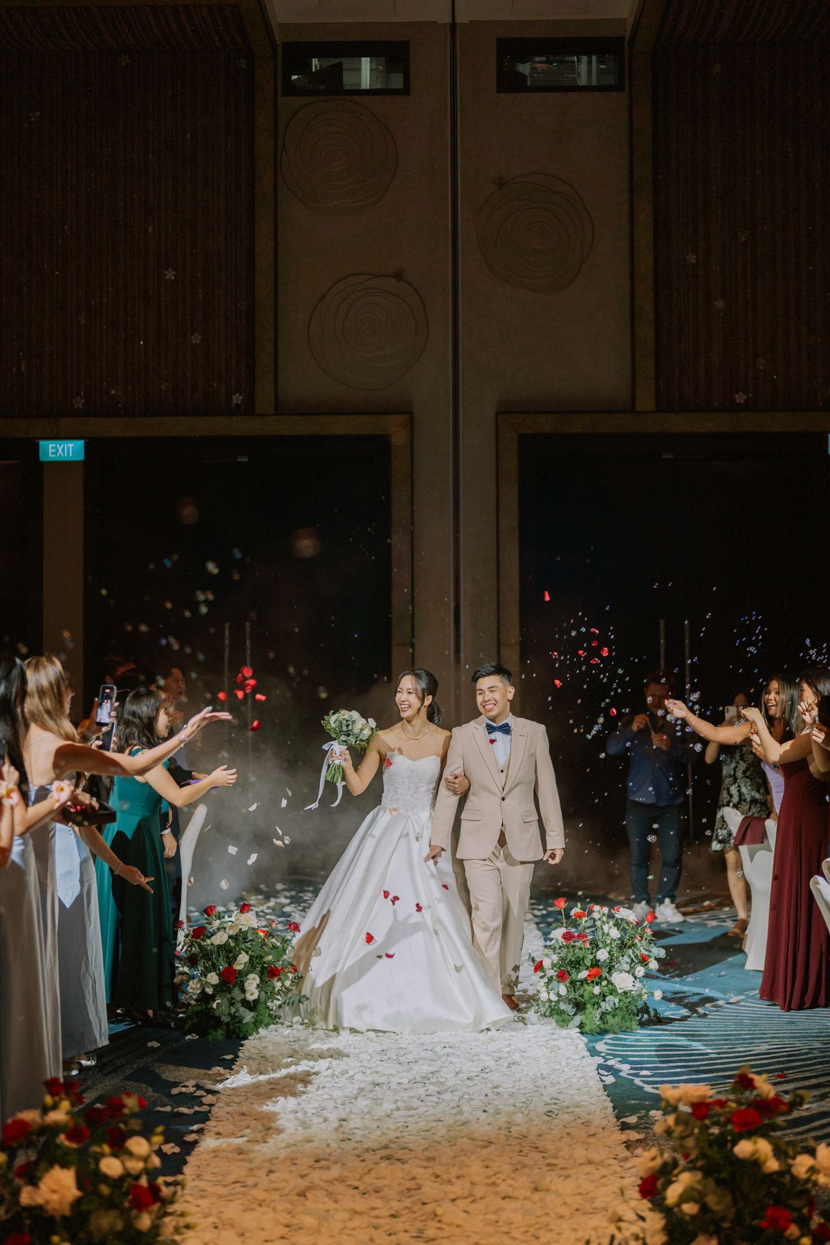 Bride and groom walk down the aisle as guests toss flower petals during an elegant indoor wedding ceremony.