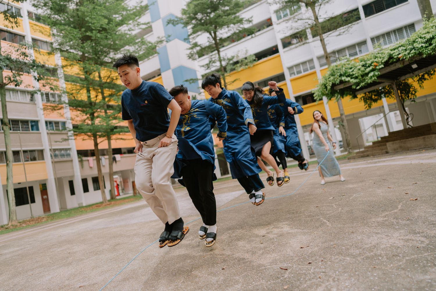 Students in blue uniforms play an outdoor jumping game in a school courtyard surrounded by colorful buildings.