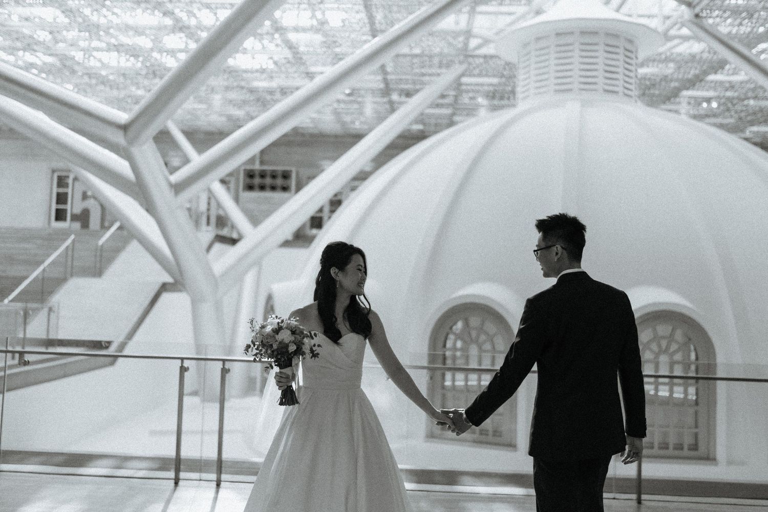 Black and white photo of a bride and groom holding hands at an elegant domed venue.