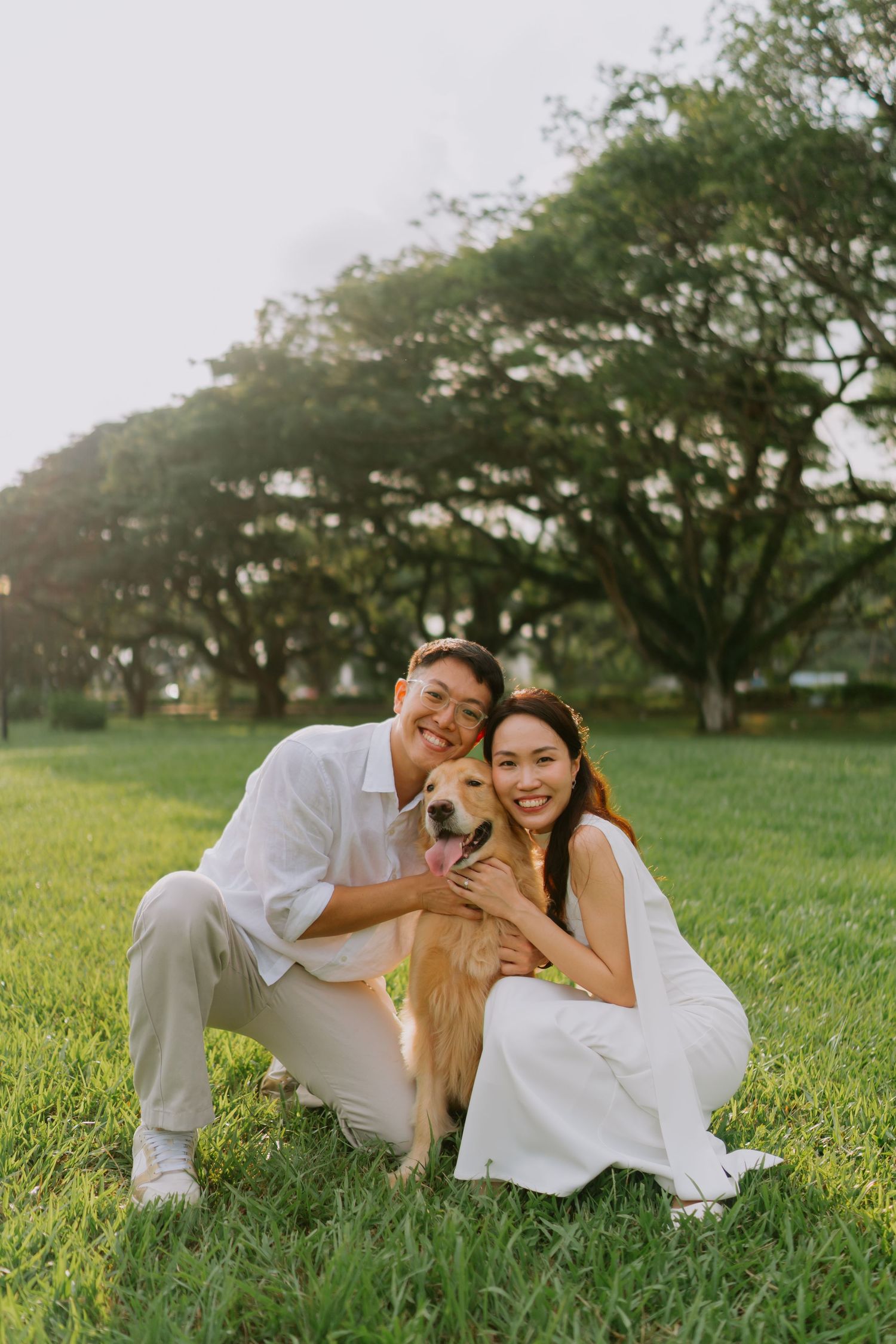 Smiling couple in white wedding clothes crouch beside golden retriever dog on sunlit green grass.