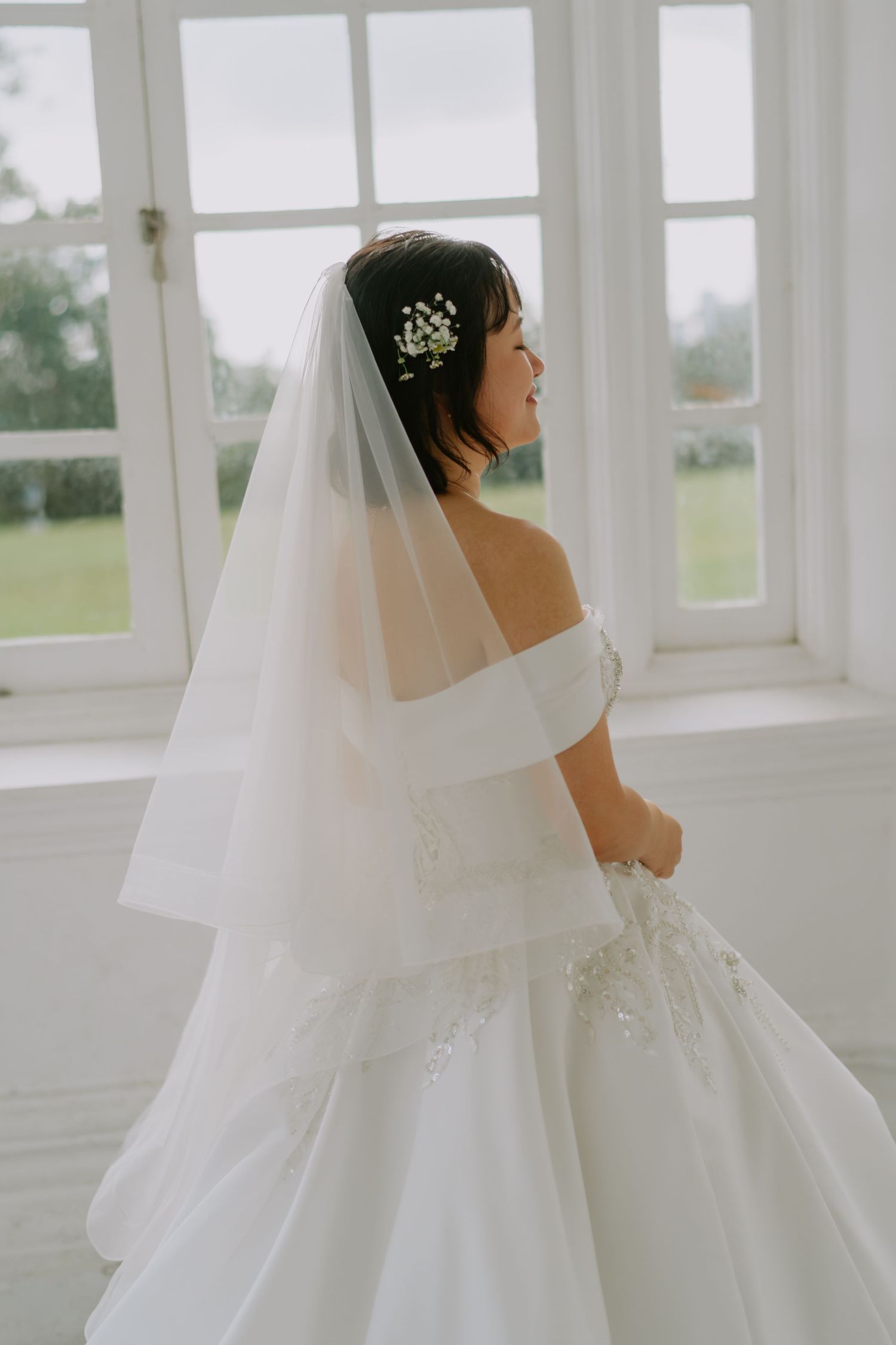 Smiling bride in an off-shoulder gown and veil standing by large windows on her wedding day.