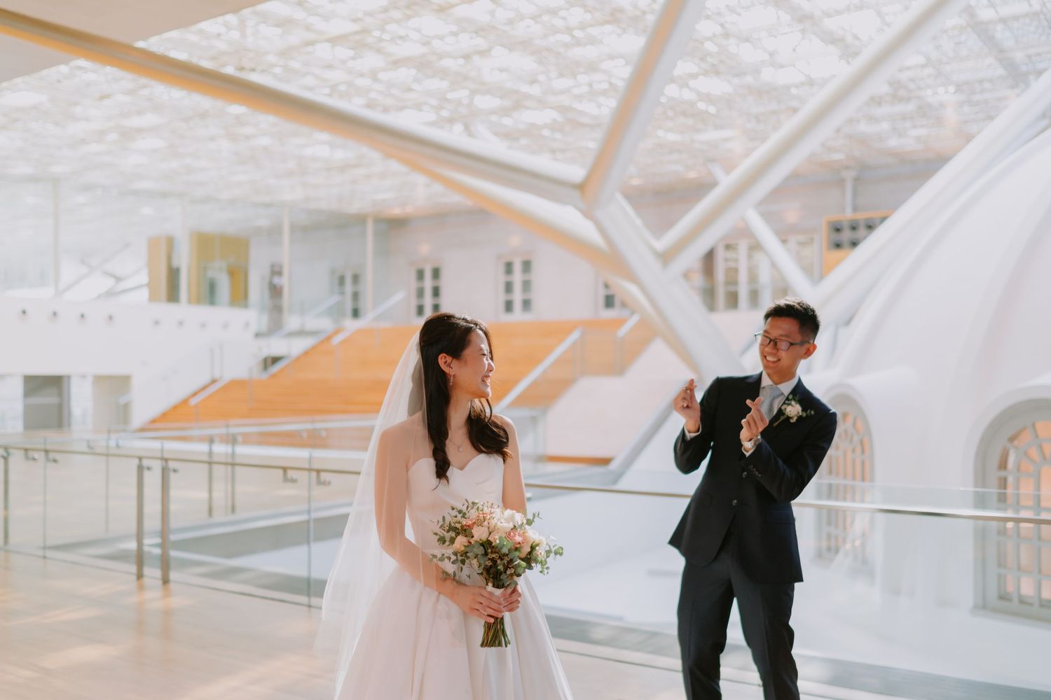 Asian bride holding bouquet laughing as groom adjusts tie in bright modern white architectural wedding venue.