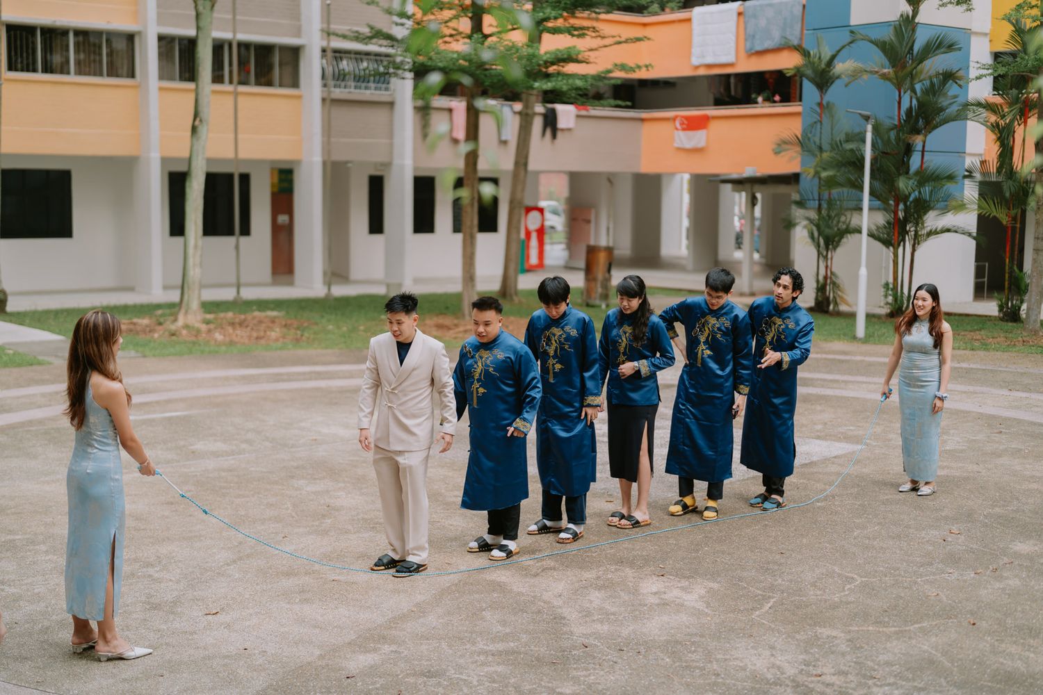 Group in blue ao dai traditional Vietnamese attire poses outdoors at a school courtyard with two people in modern outfits.