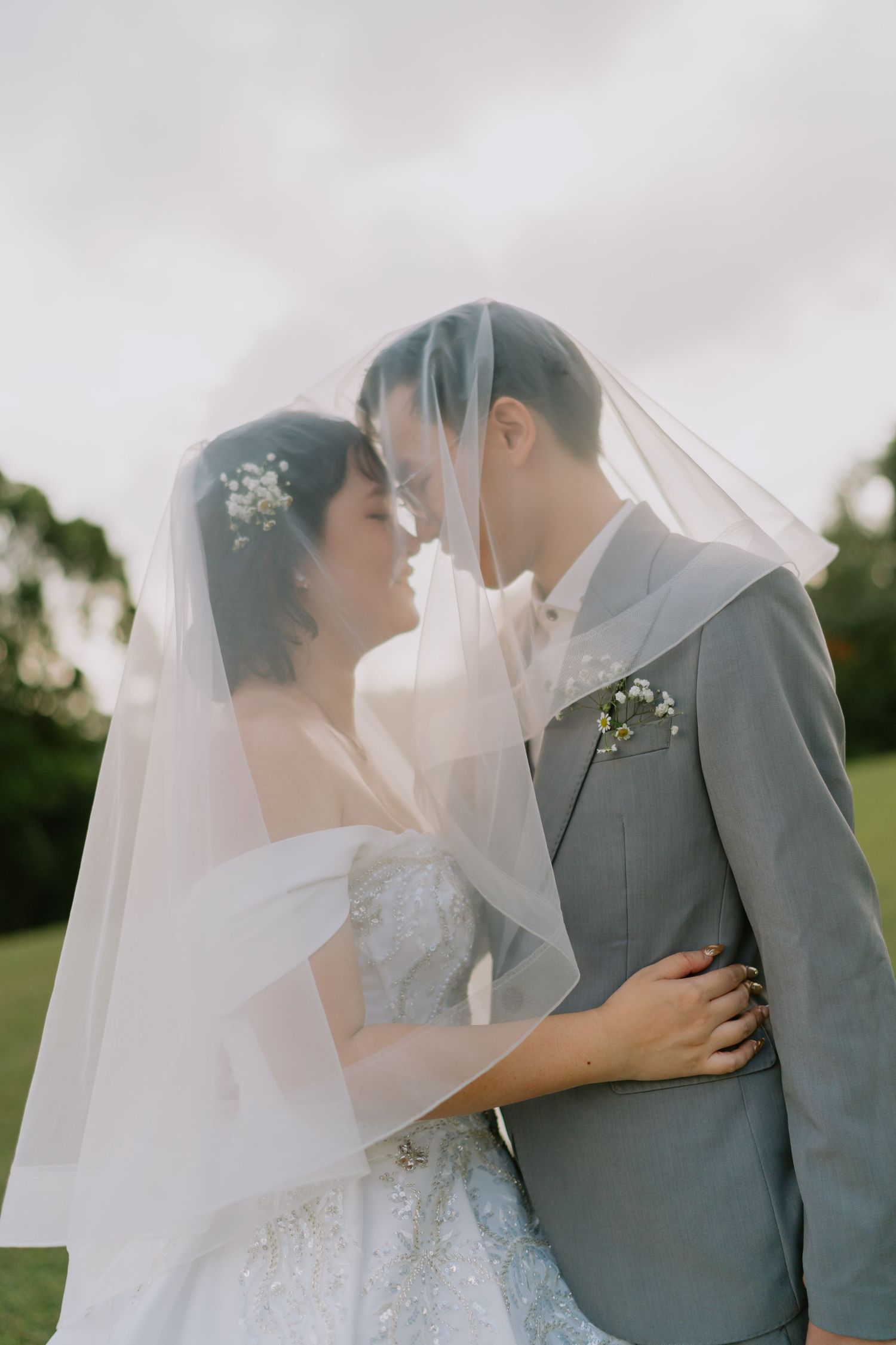 Bride and groom embrace tenderly under a sheer white veil on a lush green lawn outdoors.