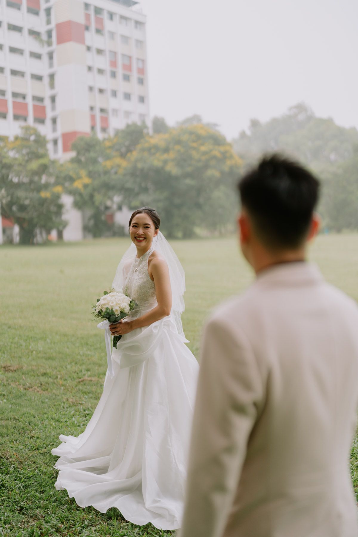 Smiling bride in white gown holding bouquet faces groom in beige suit on green lawn near building.