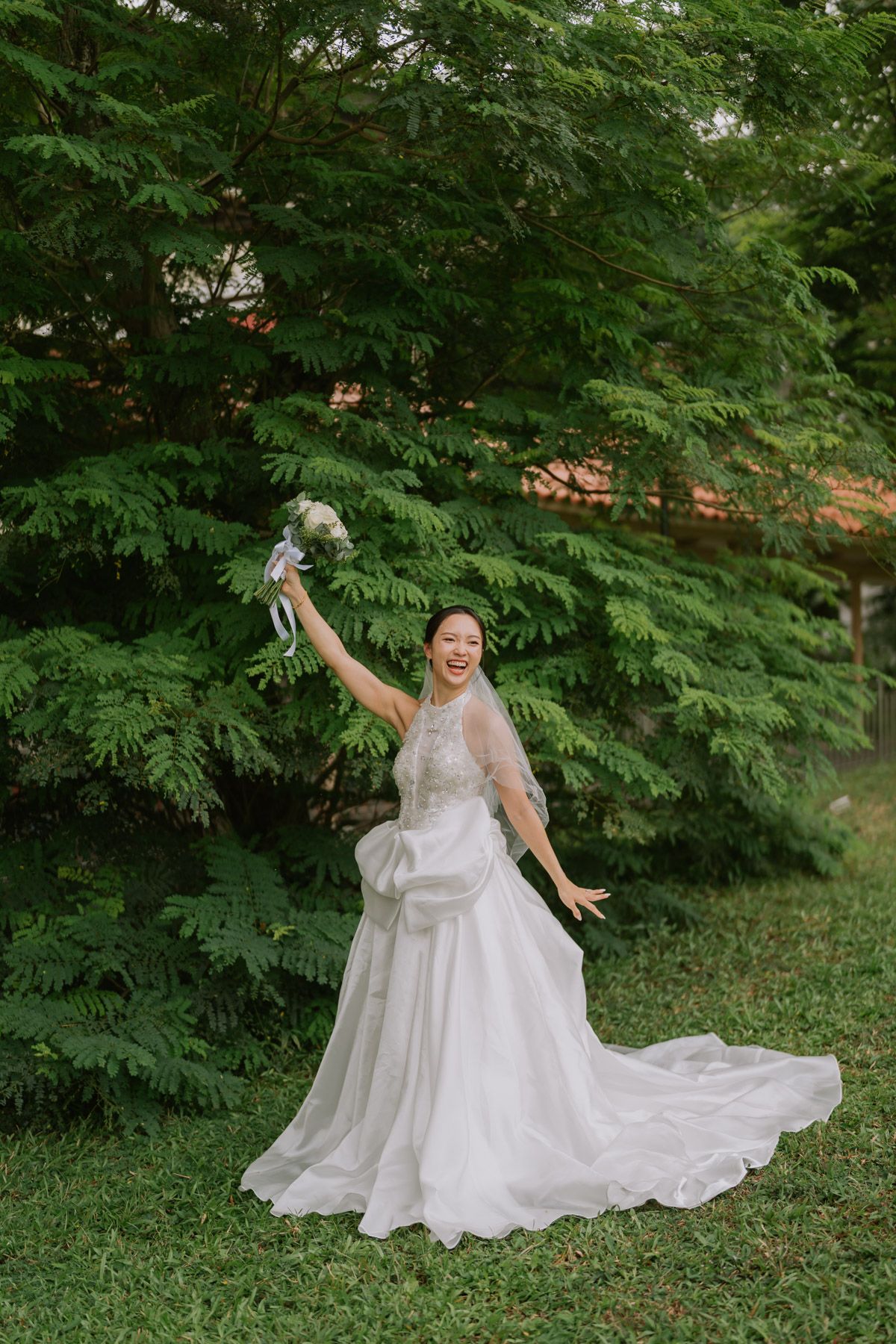 A joyful bride in a white gown raises her bouquet outdoors, smiling radiantly against lush green trees.