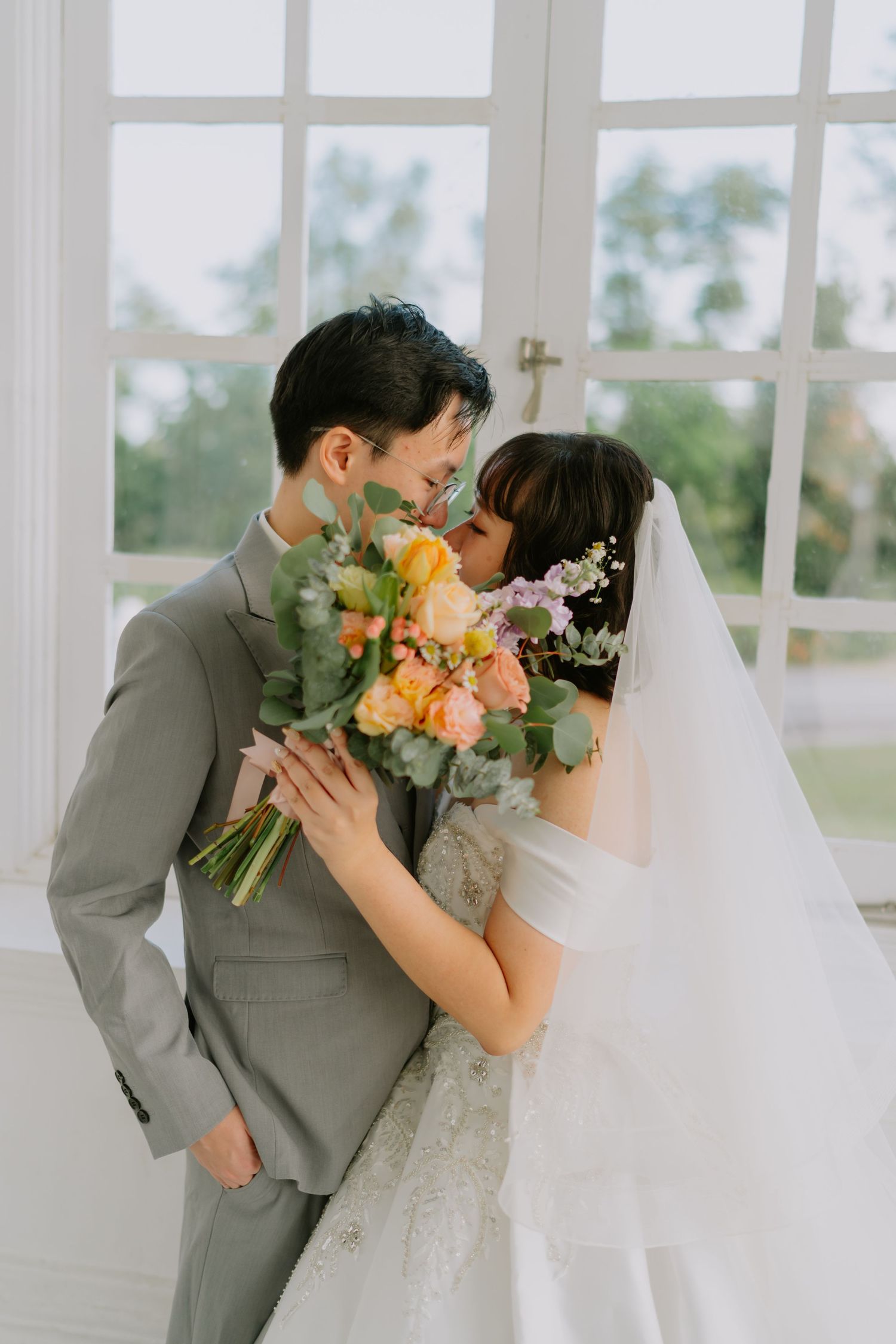 Bride and groom share a romantic moment with a colorful bouquet of flowers near bright white windows.