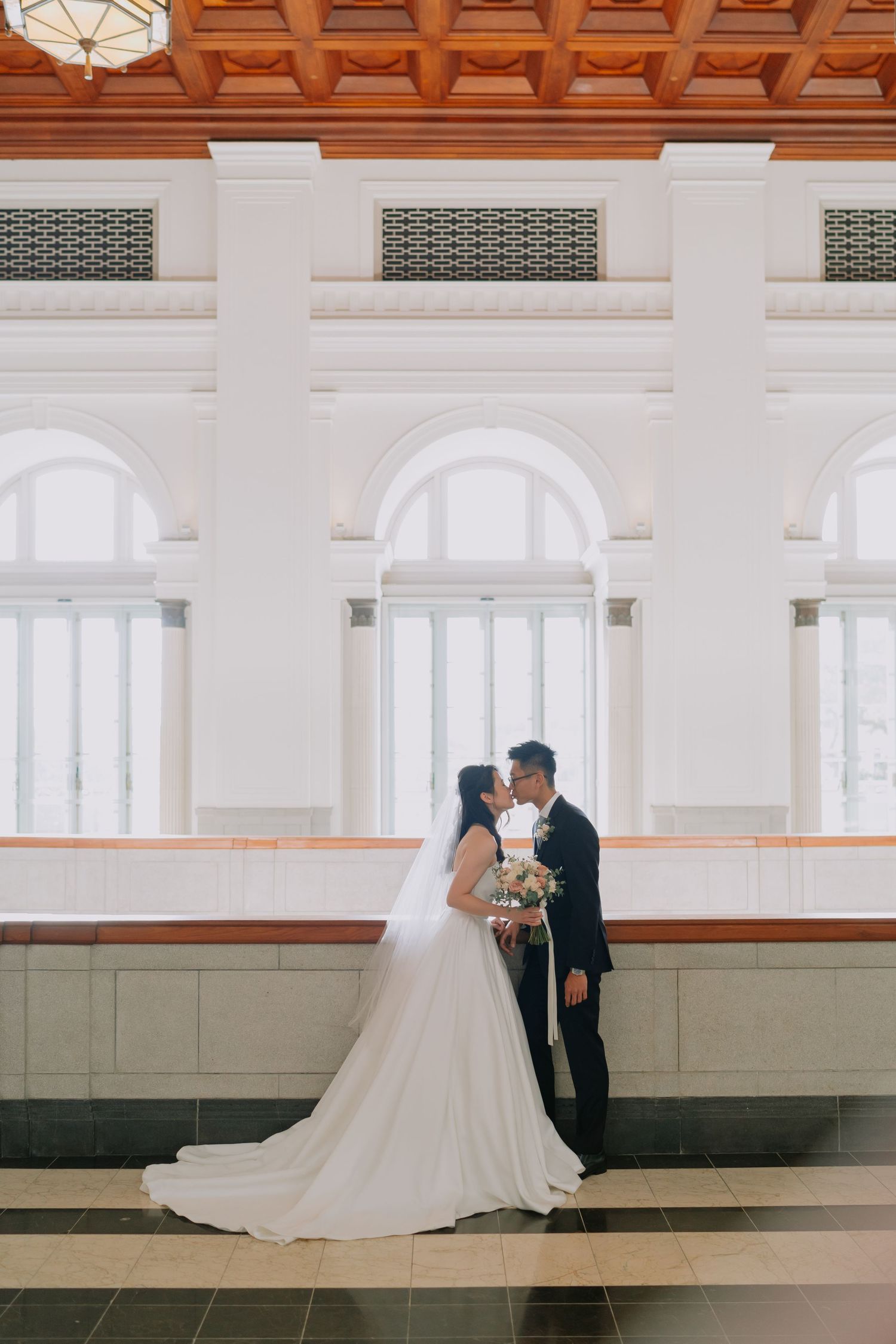 Wedding couple sharing a romantic kiss in grand white colonial hall with arched windows.