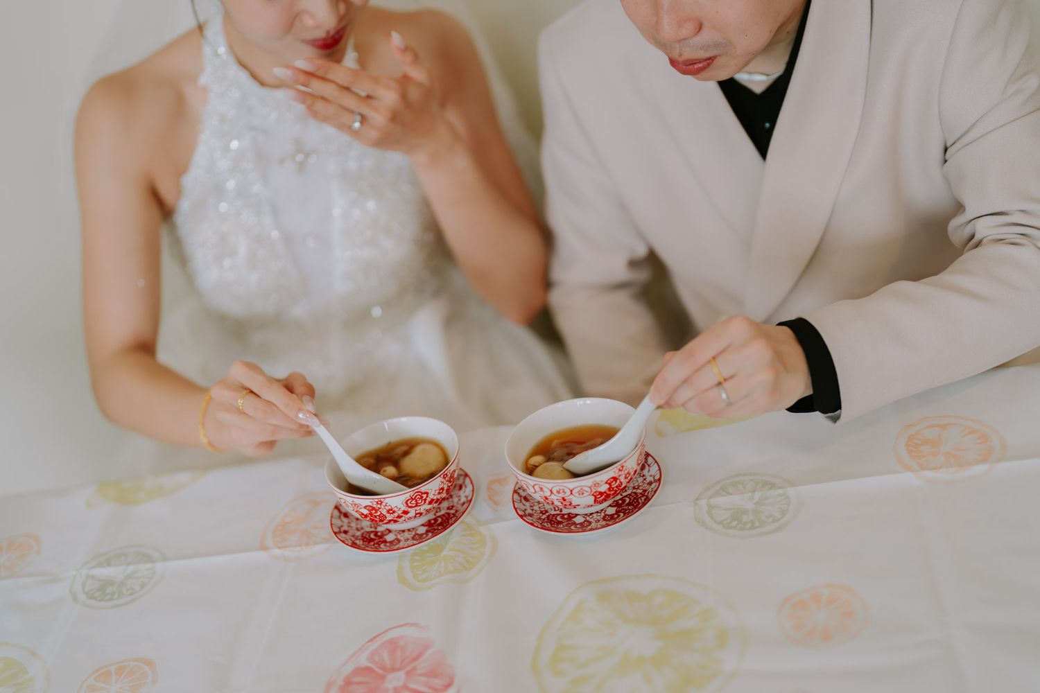 Bride and groom in wedding attire eating soup from red floral bowls with spoons at a decorated table.